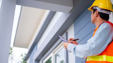 A construction inspector in a yellow hard hat and orange safety vest holds a clipboard and pen outside a modern house.