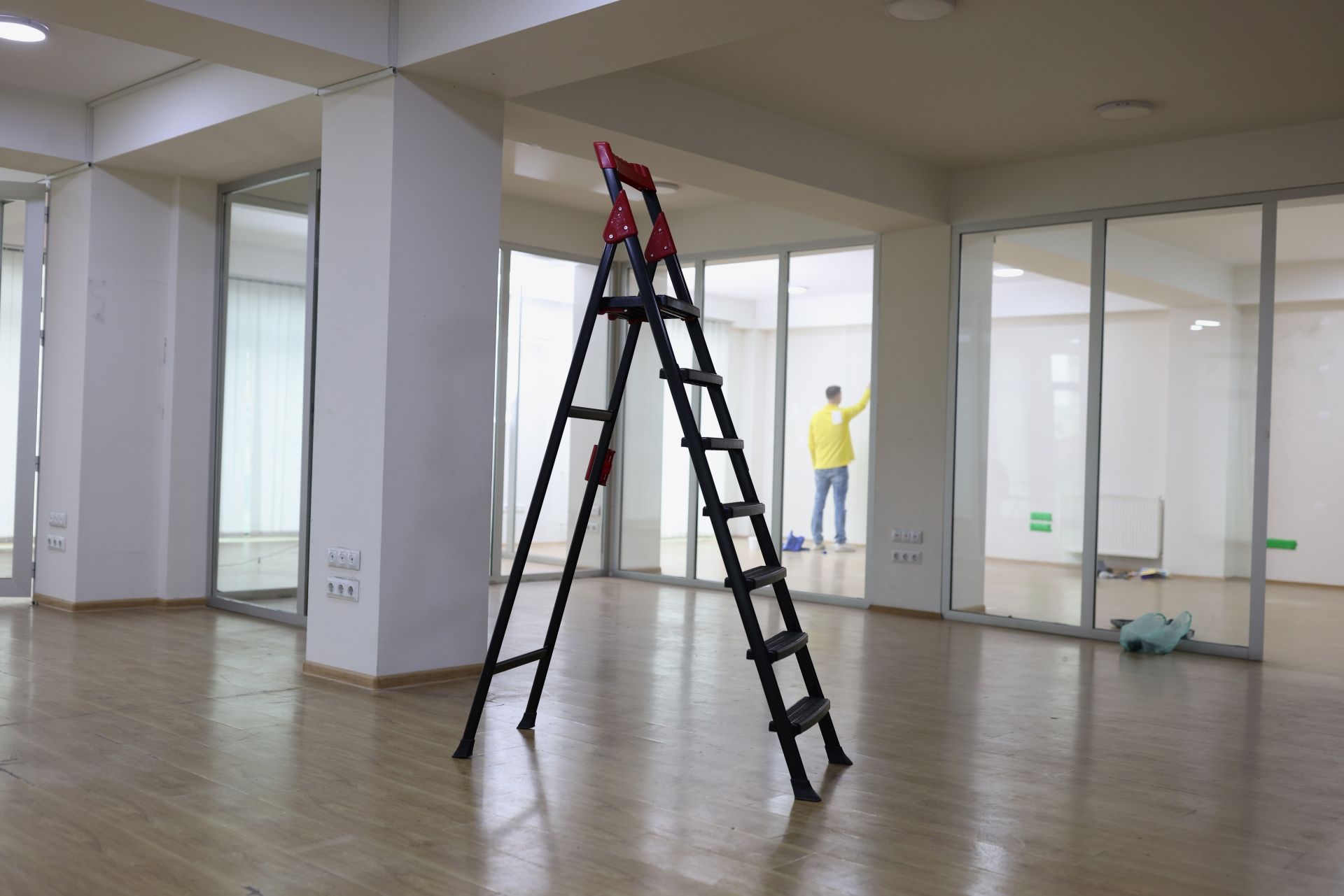 A black A-frame ladder stands in the center of an empty office with large glass windows and a person in the distance.