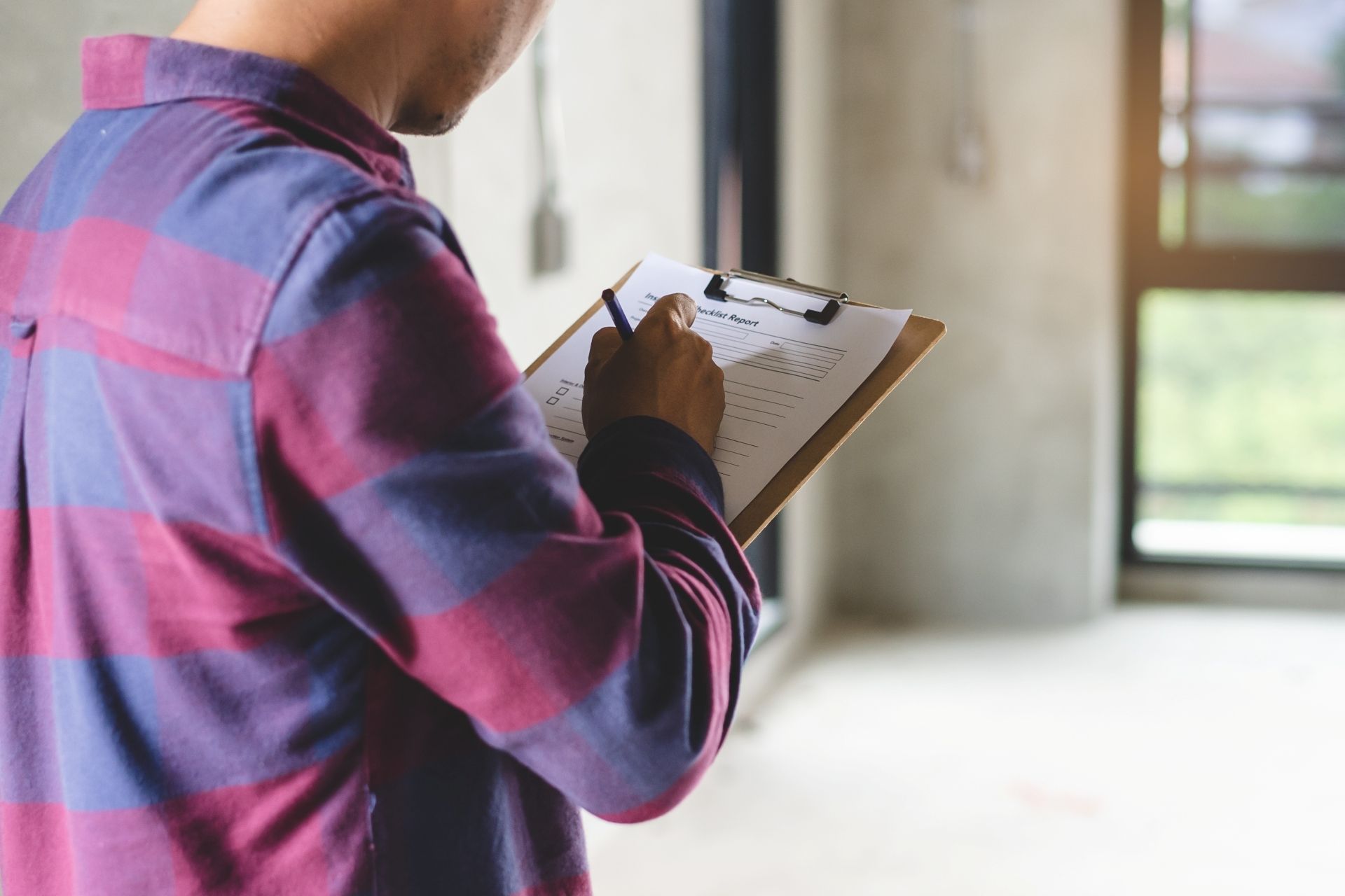 A person in a red and blue plaid shirt writes on a clipboard while standing inside an unfinished concrete room.
