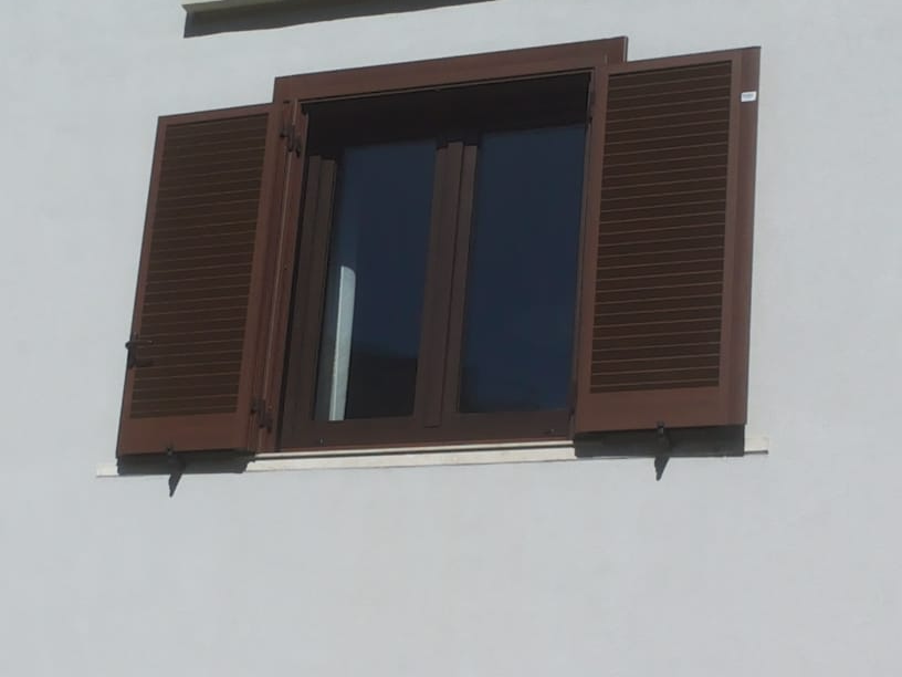 Delle persiane marroni si aprono su una finestra con cornice scura in una parete bianca.