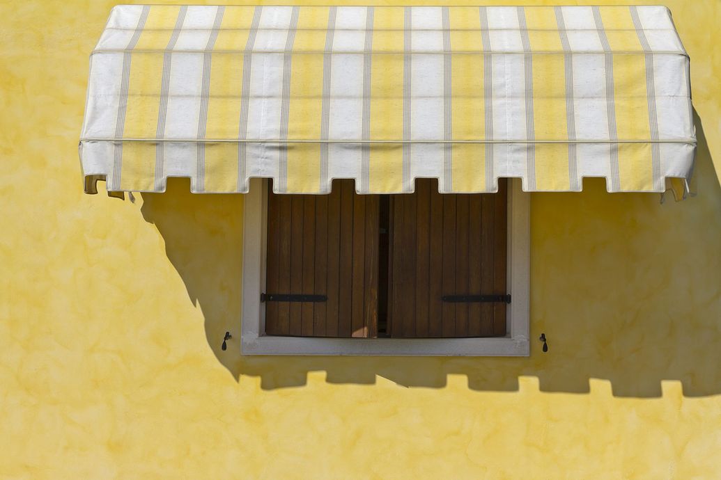 Parete gialla con finestra coperta da tenda a strisce gialle e bianche; persiane di legno chiuse.