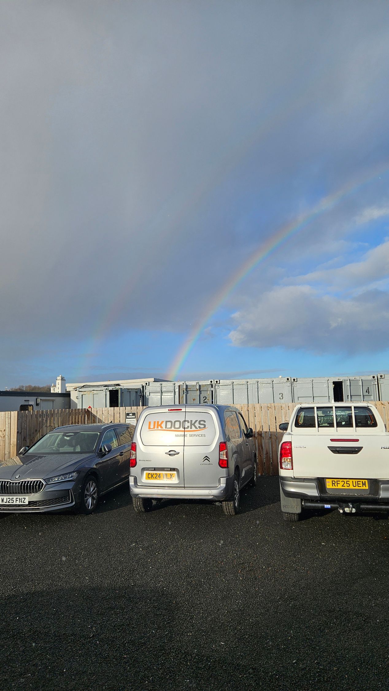 Cars parked in a gravel lot with a rainbow arching above. Gray sky, white buildings in the background.