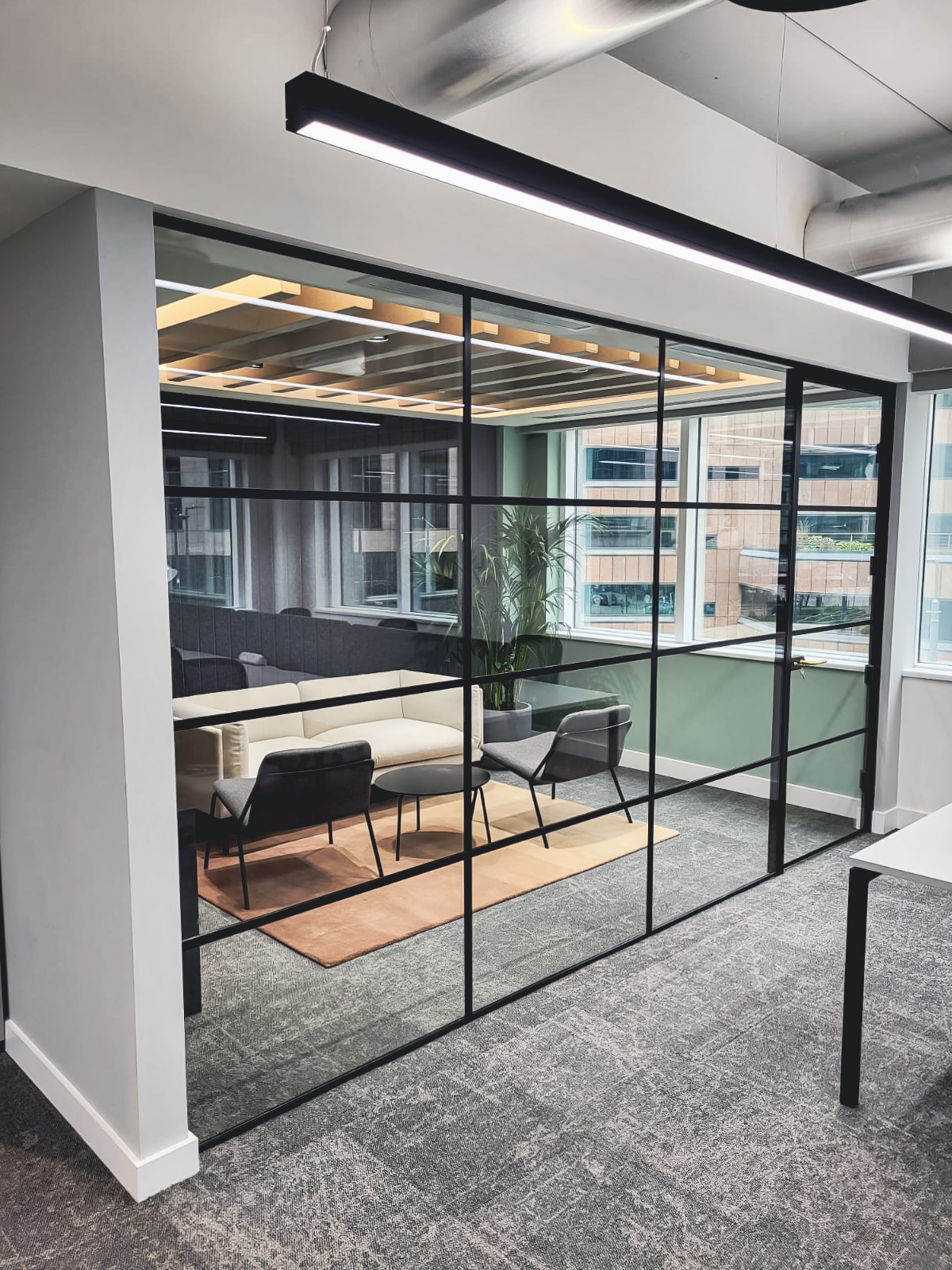 Glass-walled office space with black grid, showing seating area, neutral colors, and strip lighting.