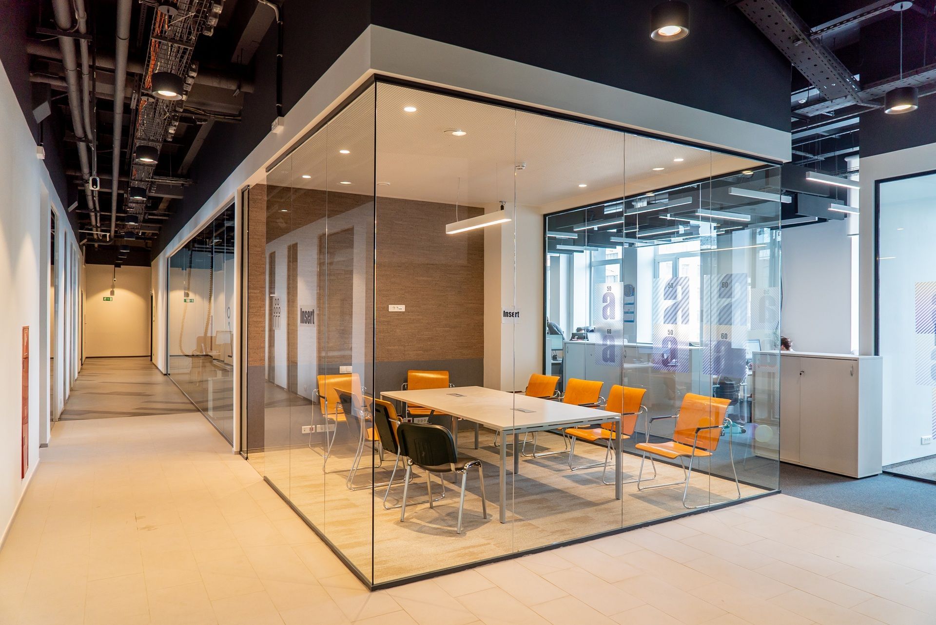 Modern office corridor with glass-walled meeting room. Orange and black chairs surround a long table.