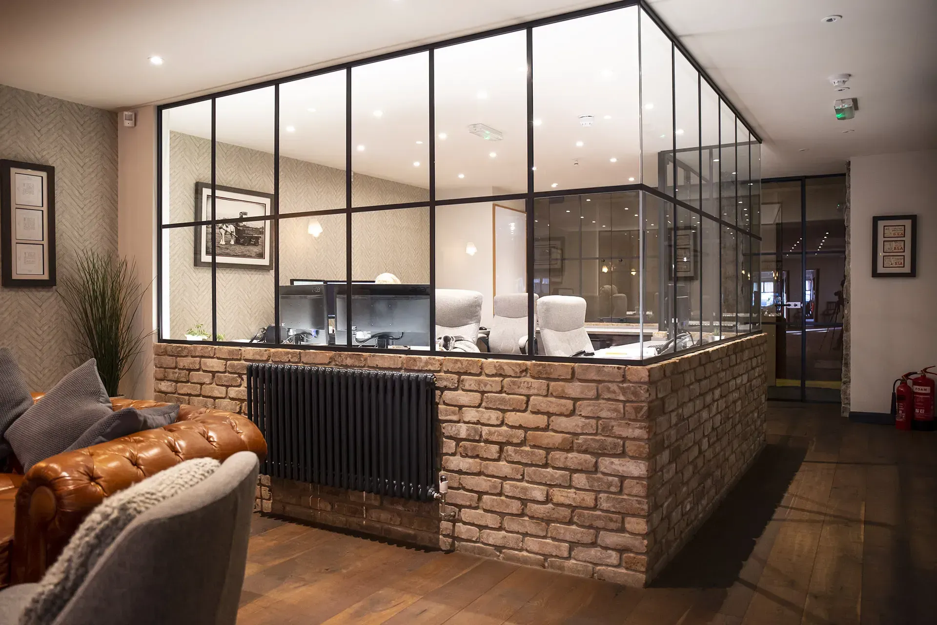 Lounge area with exposed brick wall, glass-walled office, leather sofa, and modern radiator.
