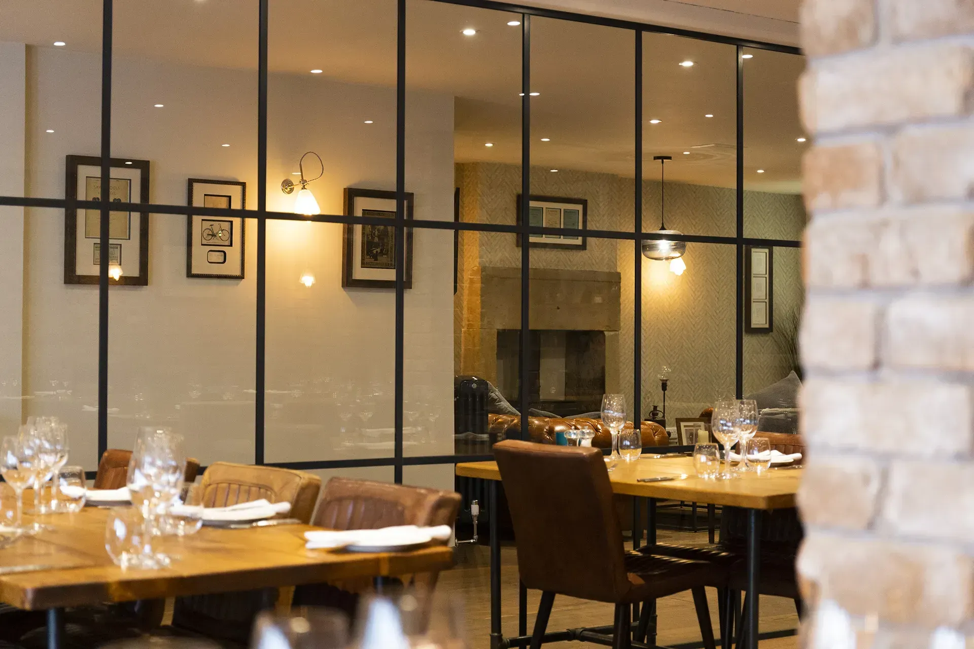 Restaurant interior with tables set for dining, visible through a glass wall. Artwork and a fireplace are in the background.