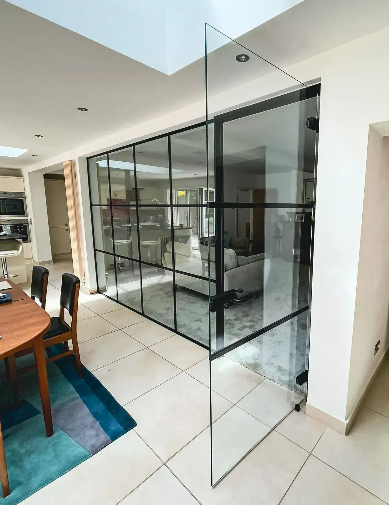 Modern interior with black-framed glass wall and open glass door. Dining room with table and rug.