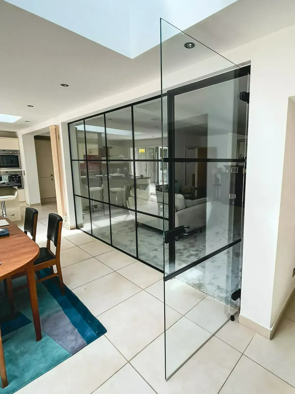 Interior view: room with a partially open, black-framed glass wall. A dining area and living space are visible.
