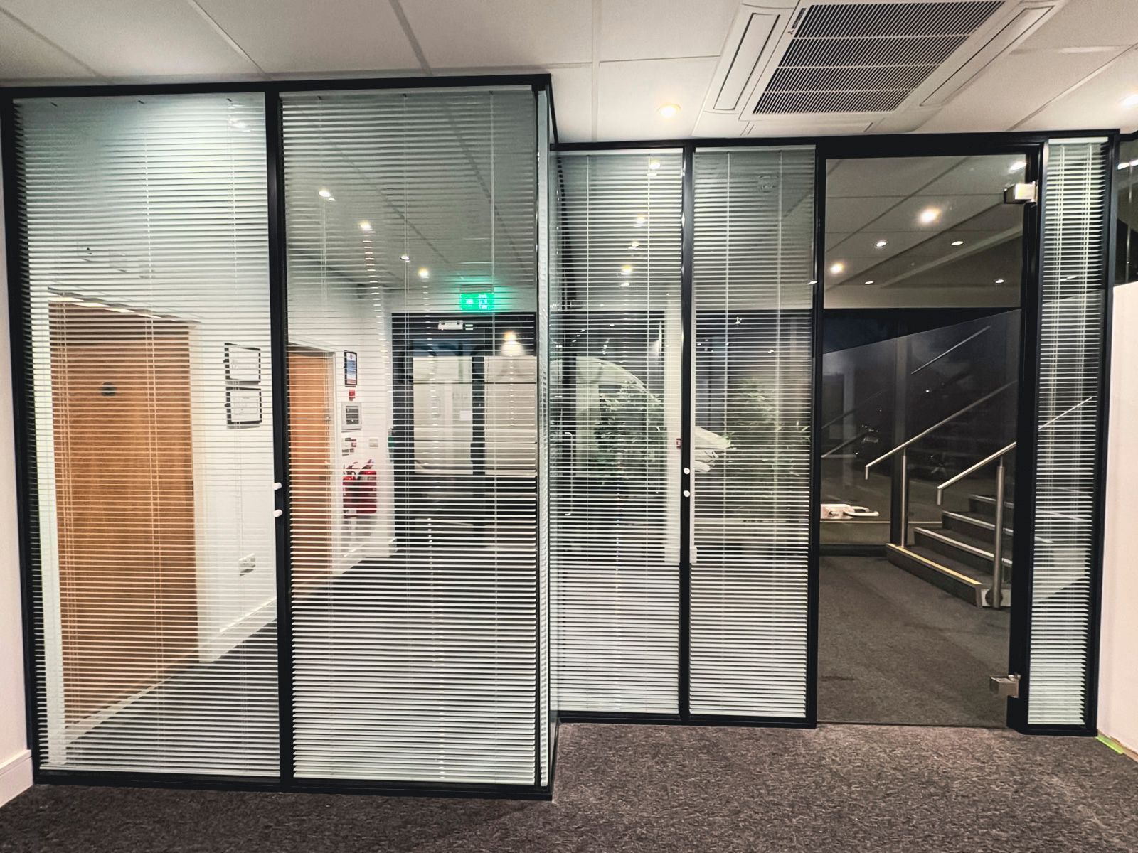 Glass office partitions with horizontal blinds. Black frames, gray carpet, and interior view with exit sign.