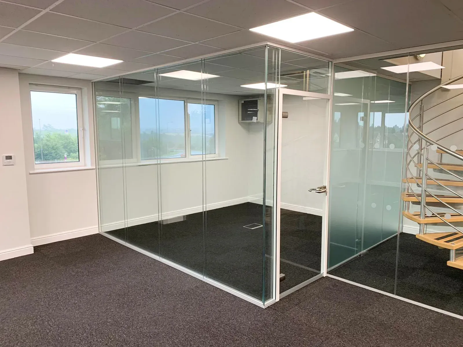 Office space with glass-walled room, windows, spiral staircase, and dark carpet.