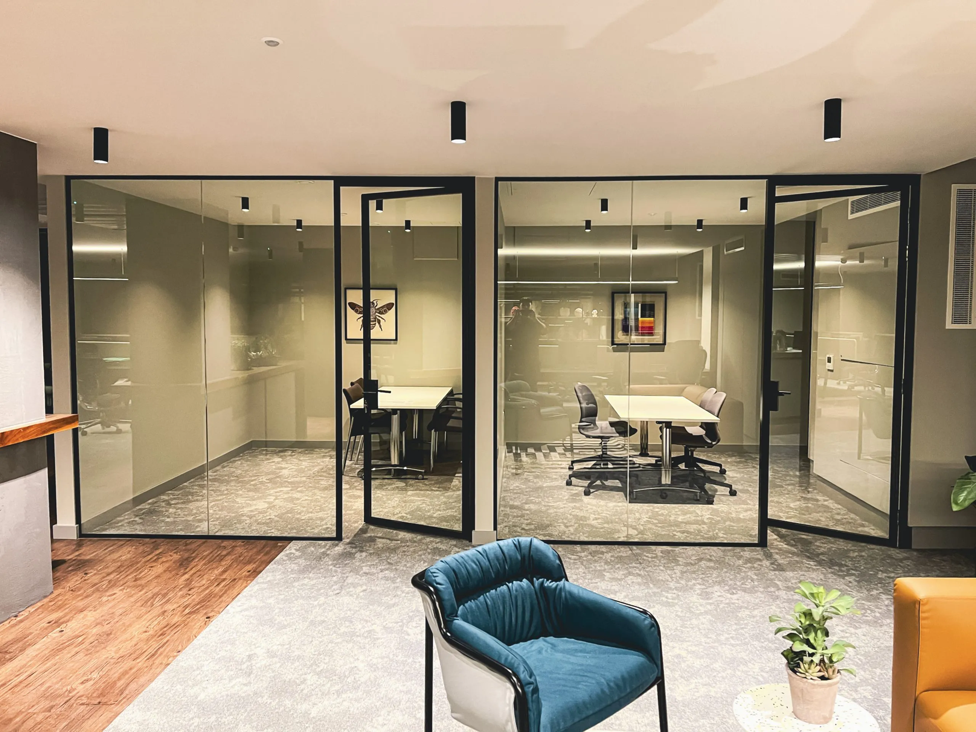 Office with glass-walled meeting rooms, furnished with tables and chairs. A blue armchair and plants are in the foreground.