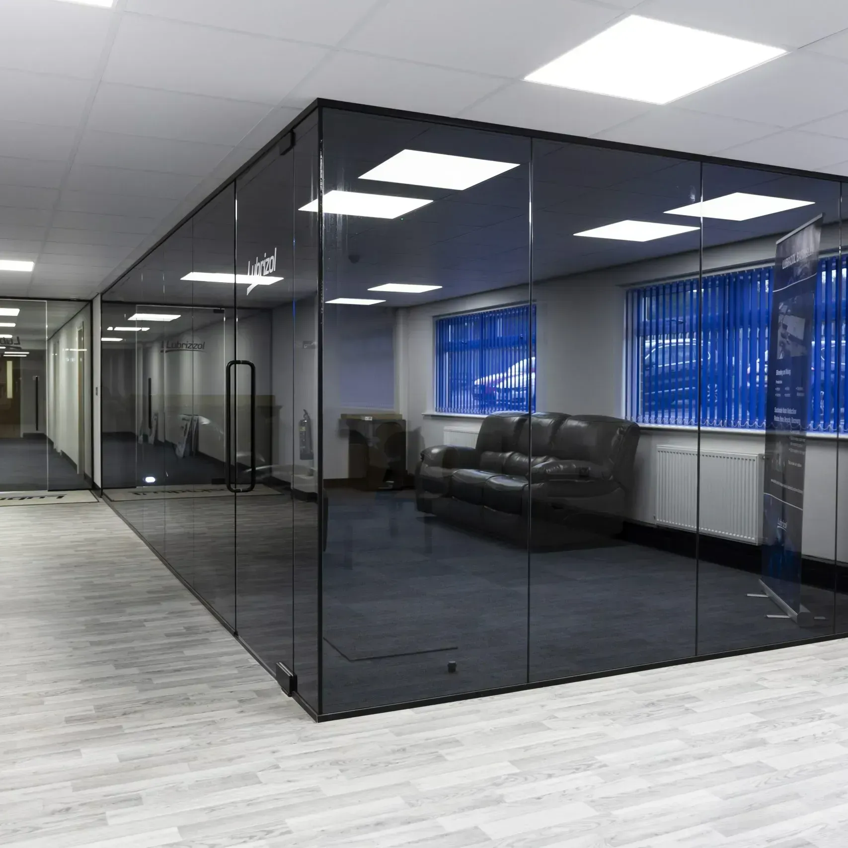 A glass-walled office with a black leather sofa, blue blinds, and fluorescent lights.