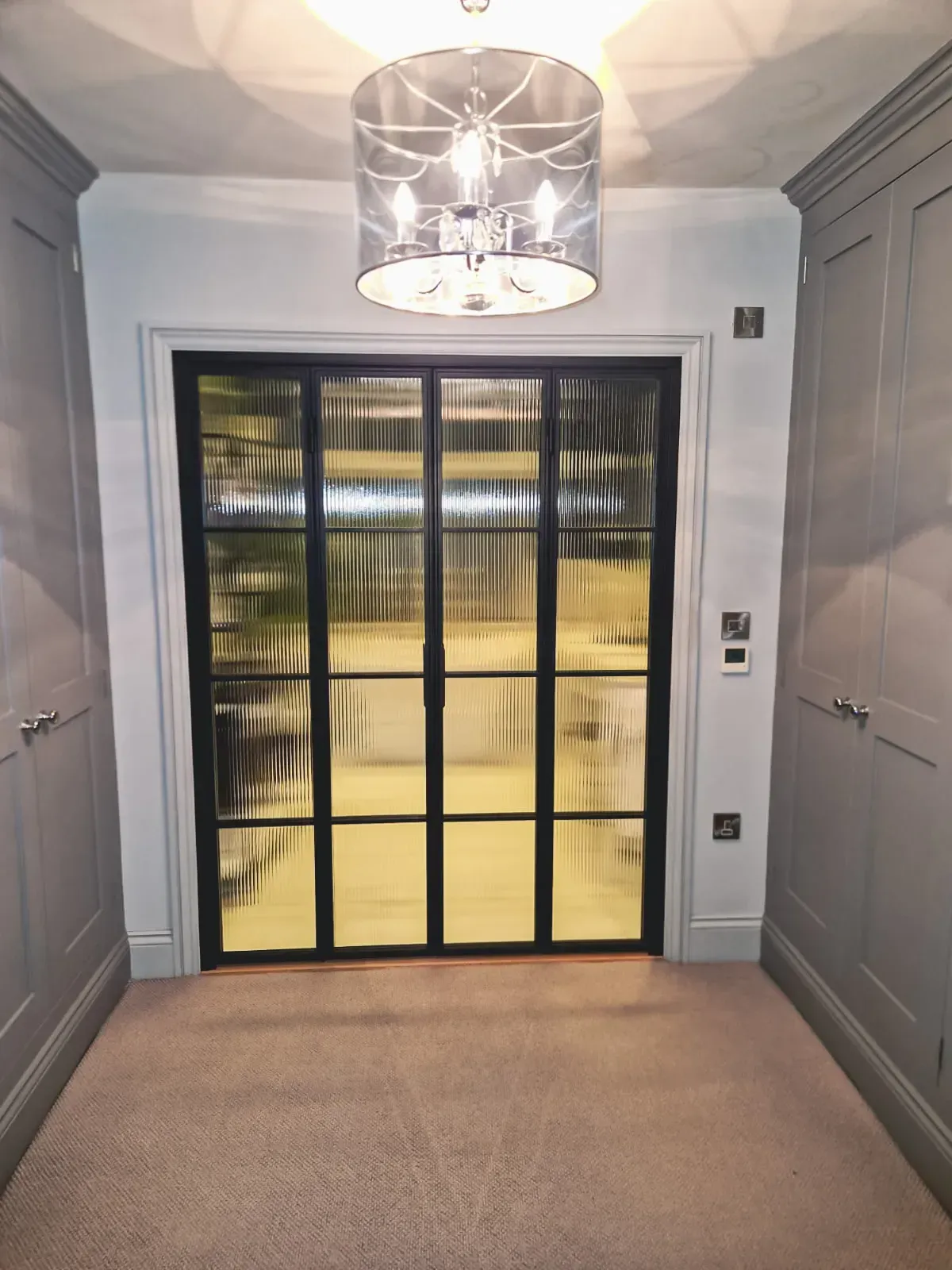 Walk-in closet with gray cabinets, black-framed glass door, beige carpet, and a light fixture.