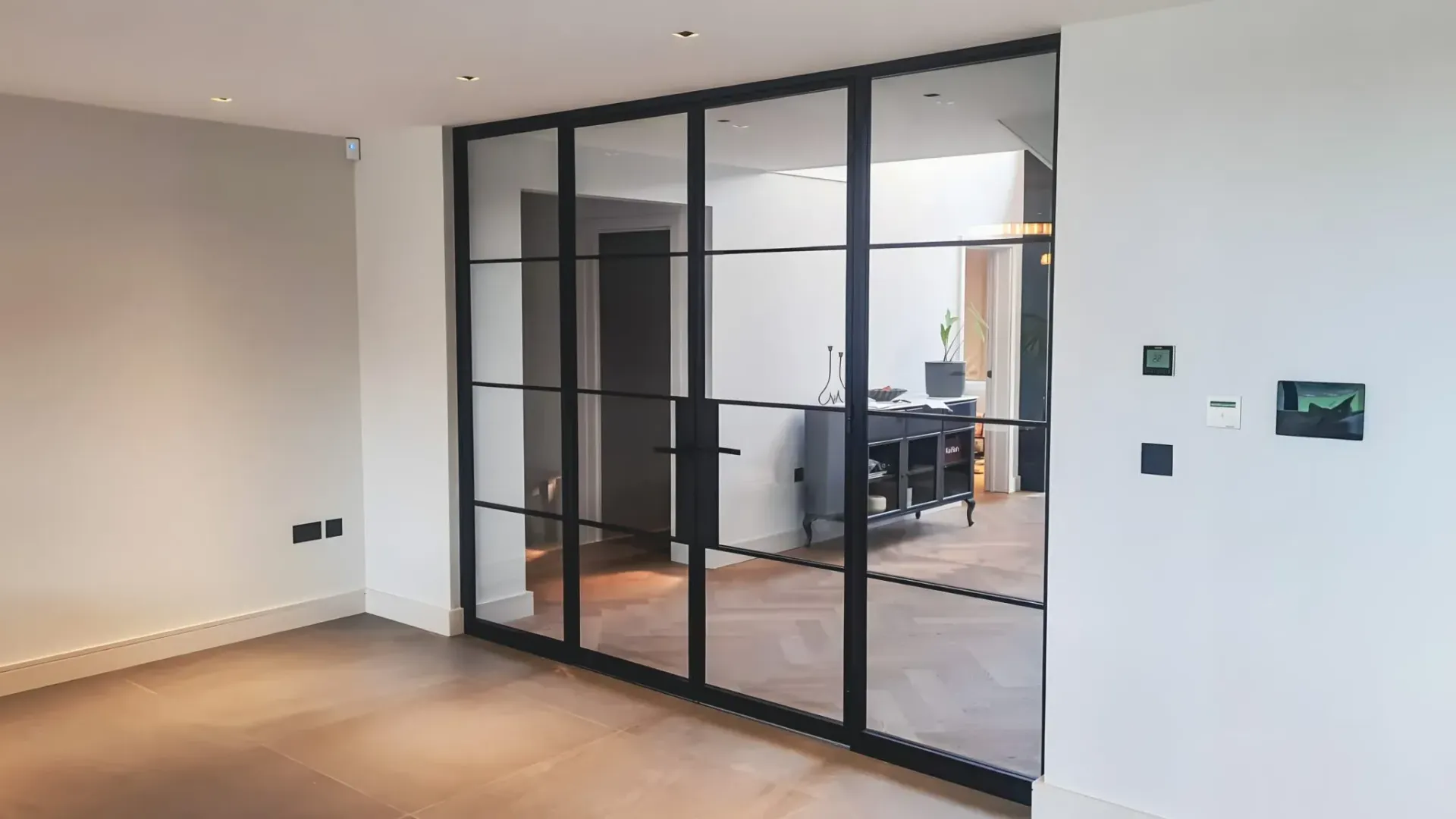 Black-framed glass doors separate two rooms. One room has a desk, the other is empty with wood floors.