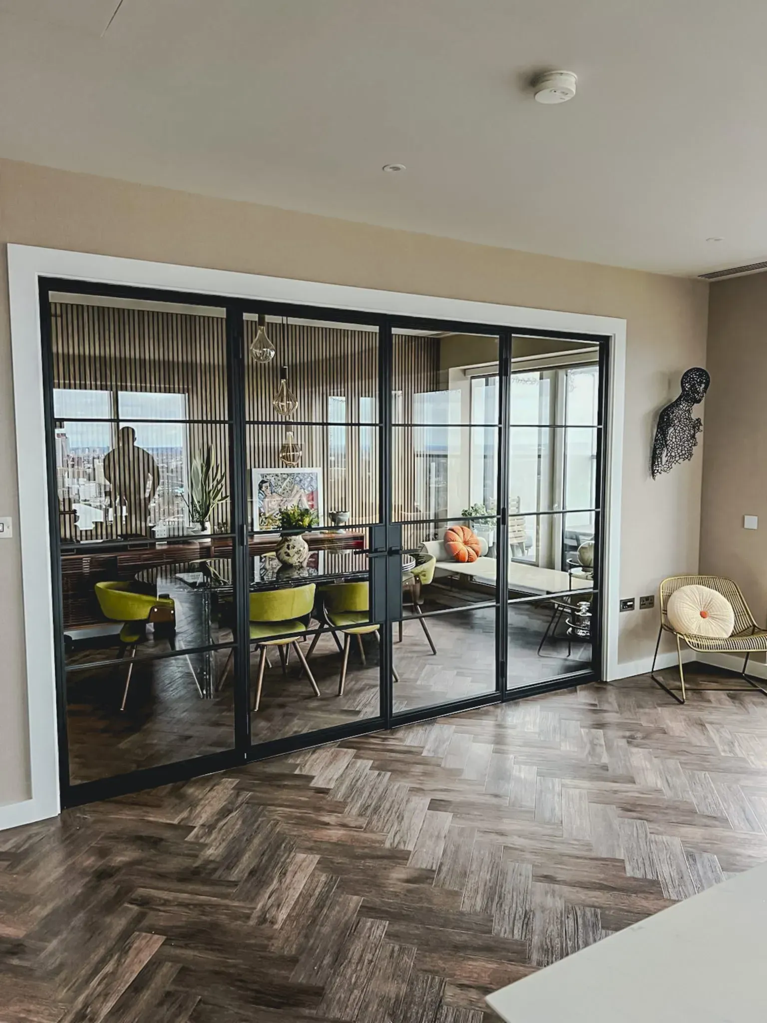 Glass-panelled doors with black frames separate two rooms. Brown herringbone floor and yellow chairs are visible.