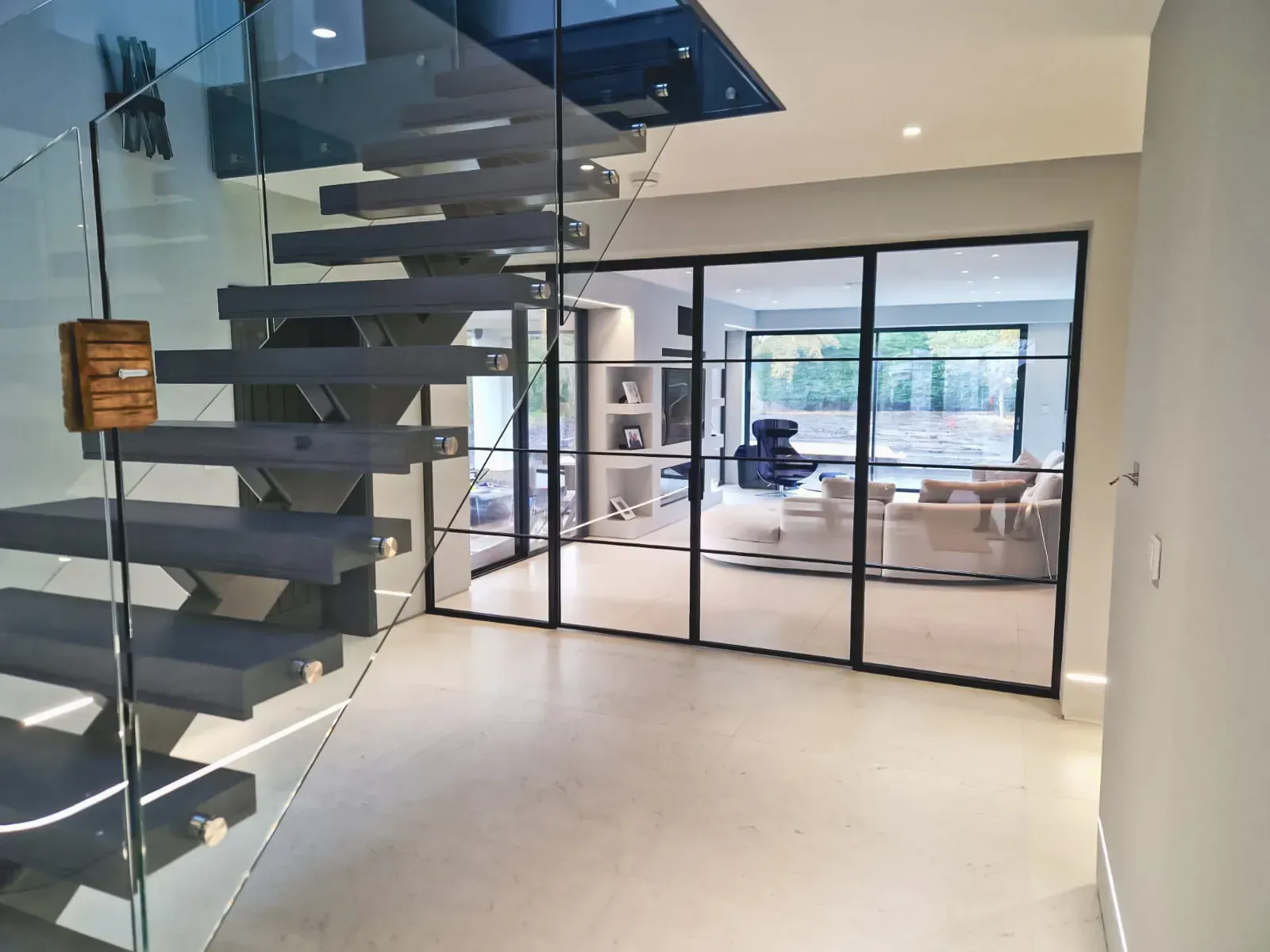 Modern interior: floating staircase, glass and black framed doors, leads to a living room with couches.