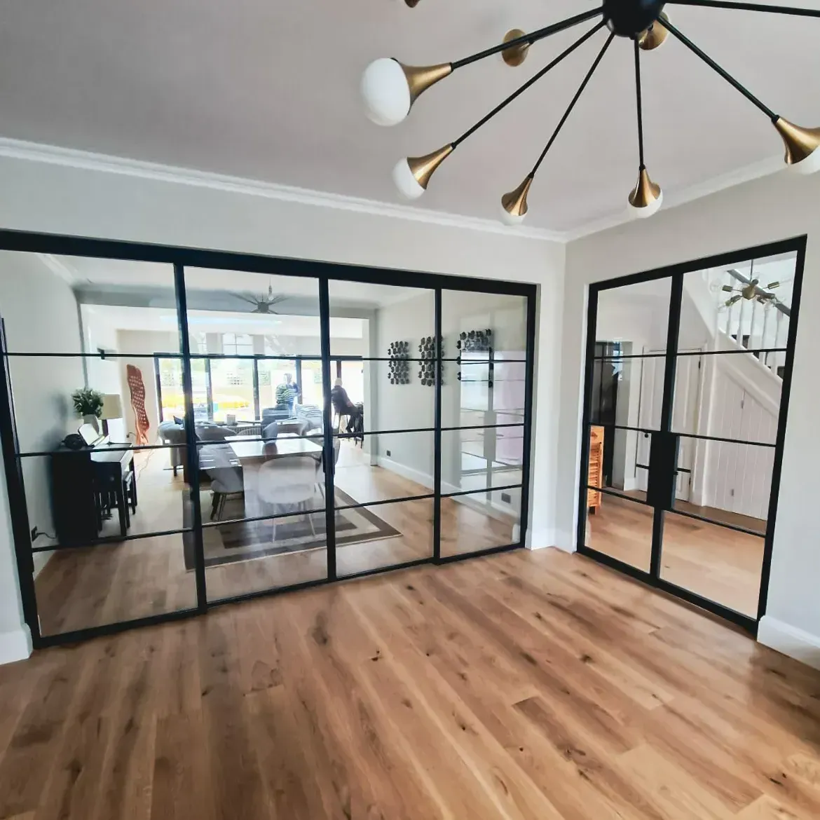 Interior with black framed glass doors, wooden floors, chandelier. View into a living room.