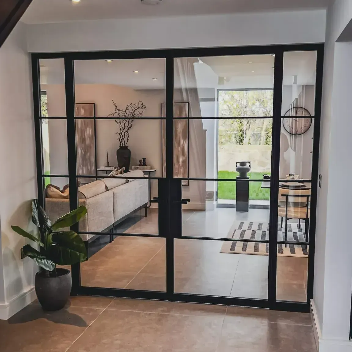 Black-framed glass doors separate two rooms. A living room with a couch and plants is visible through the doors.