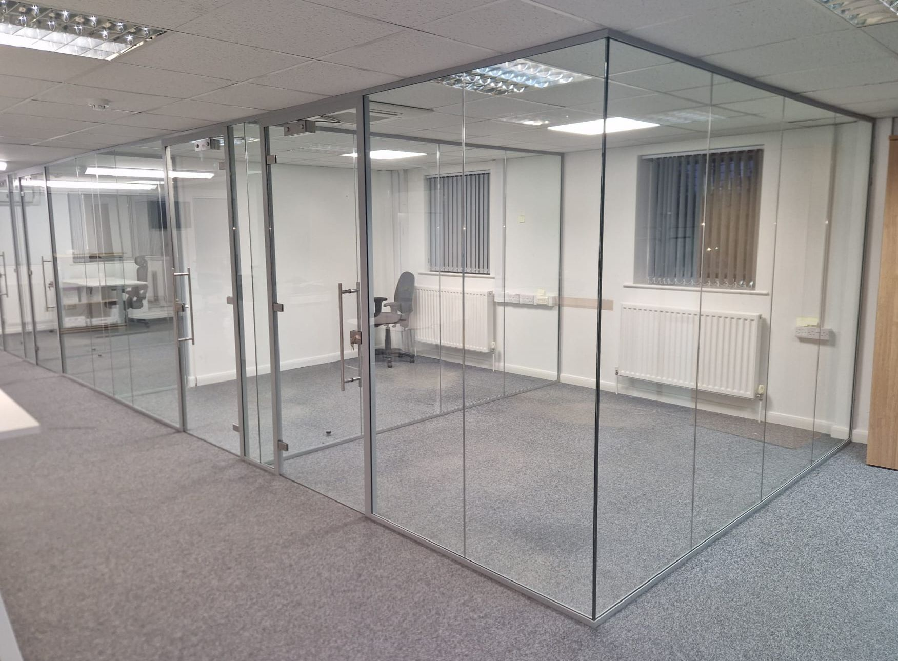 Office space enclosed by glass walls, gray carpet, white walls, and fluorescent lights.