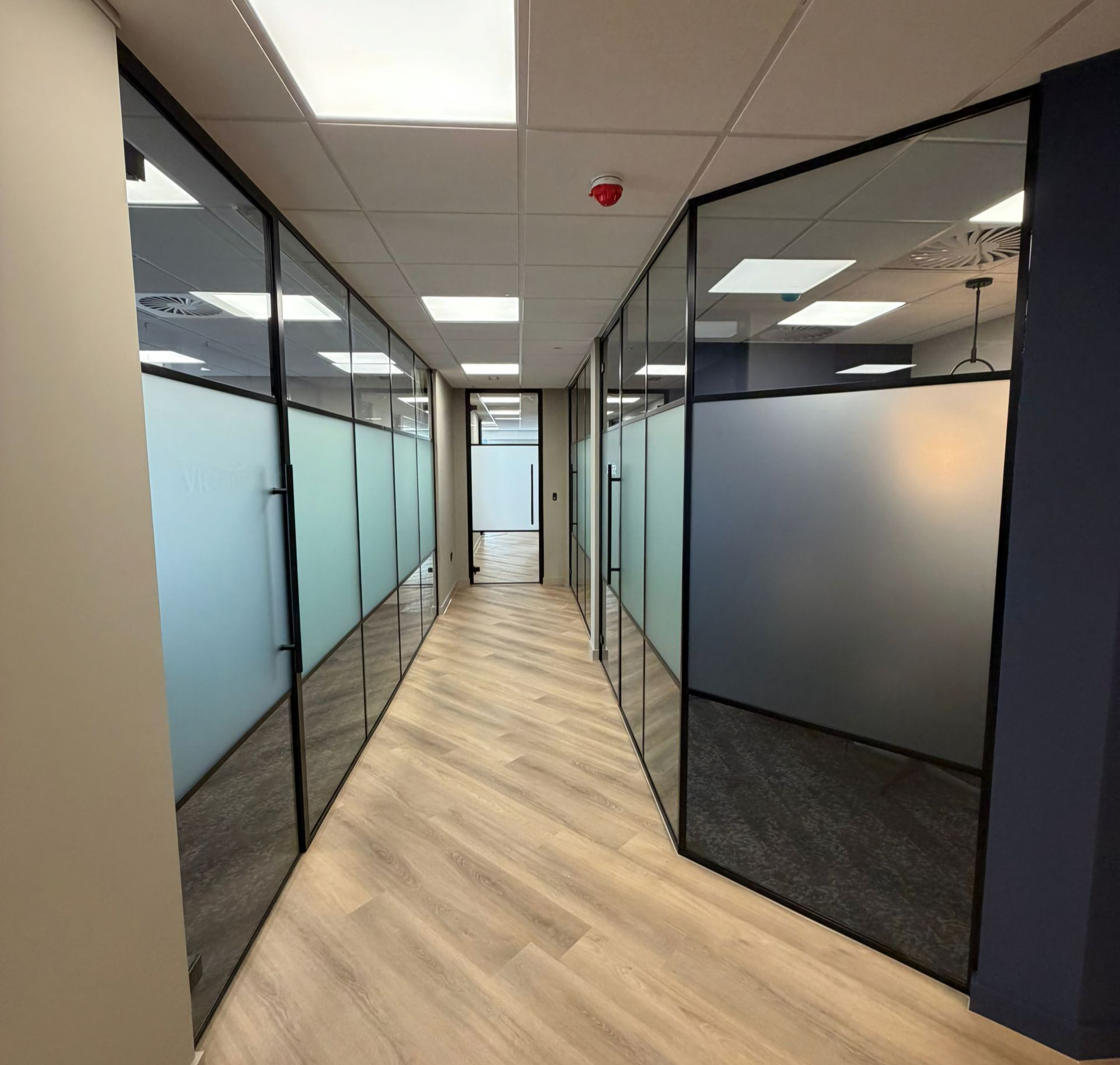 Office hallway with glass-walled rooms, frosted glass panels, wood-look flooring, and recessed ceiling lights.