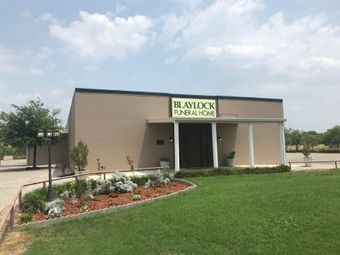 Small beige funeral home building with sign, front porch, landscaping, and green lawn under a cloudy sky
