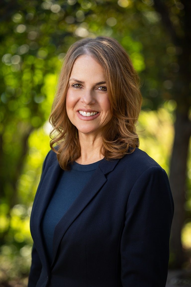 Woman in navy blazer smiles, outdoors, soft focus greenery background.