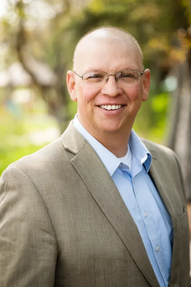 Bald man with glasses wearing a light-colored blazer and a blue shirt, smiling outdoors.