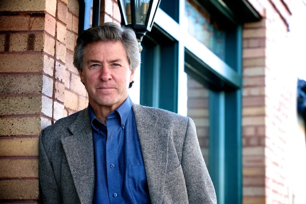 Man in blazer leans against brick wall and teal window frame, looking at the camera.