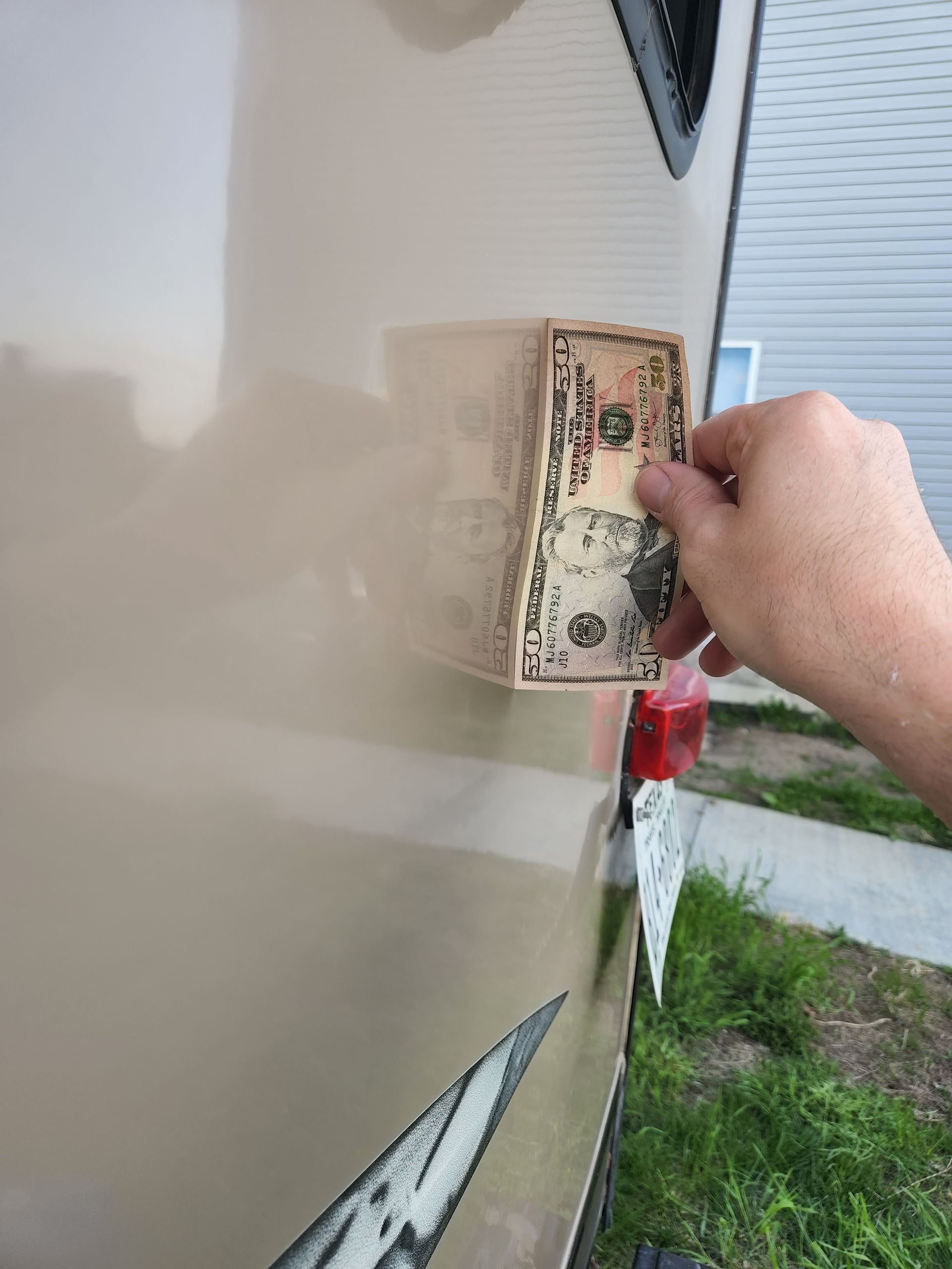 Hand cleaning a beige RV side panel with a dollar bill on a grassy driveway.