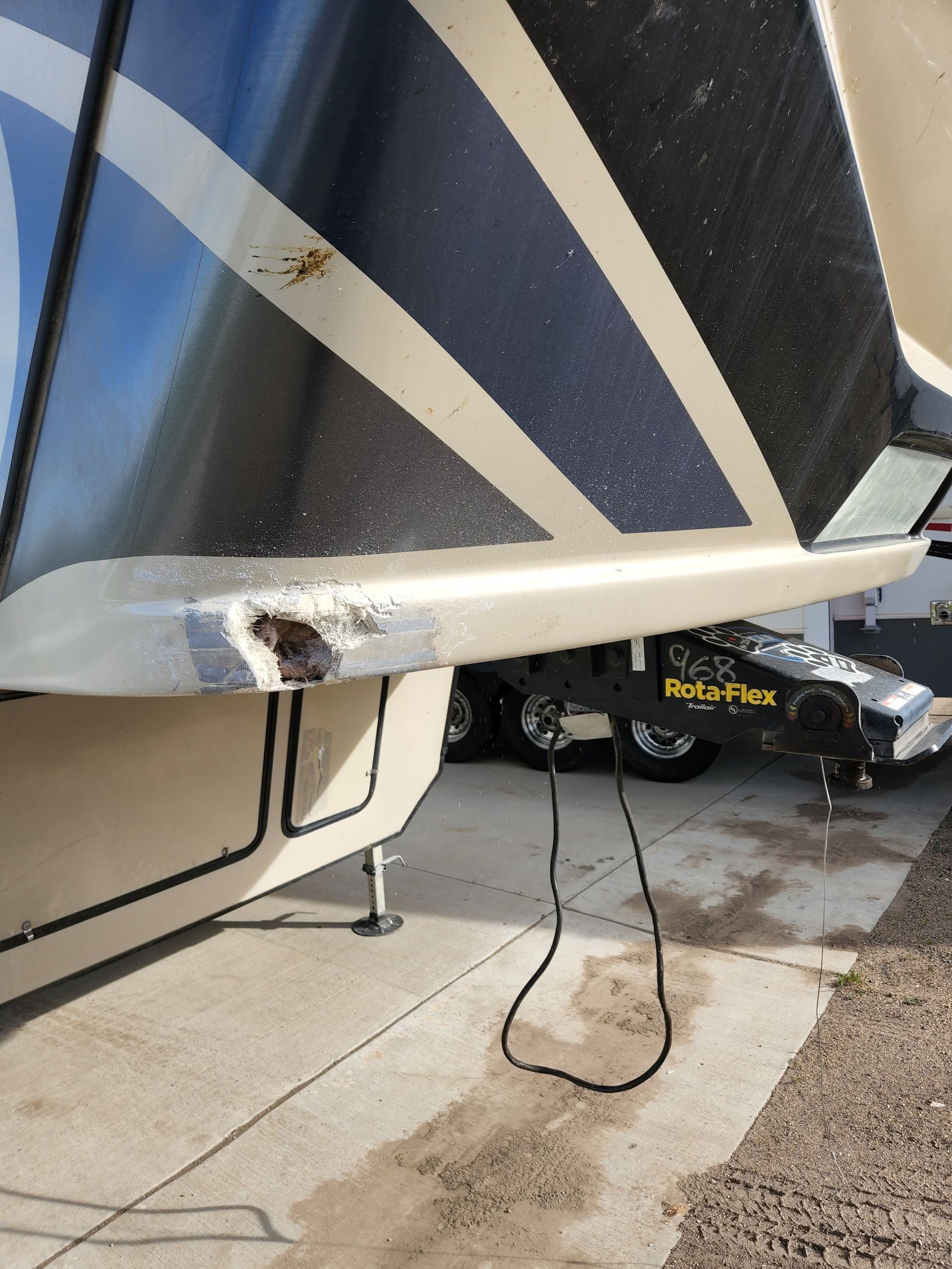 Close-up of a damaged RV corner with a torn white trim piece and hanging cable near the driveway.
