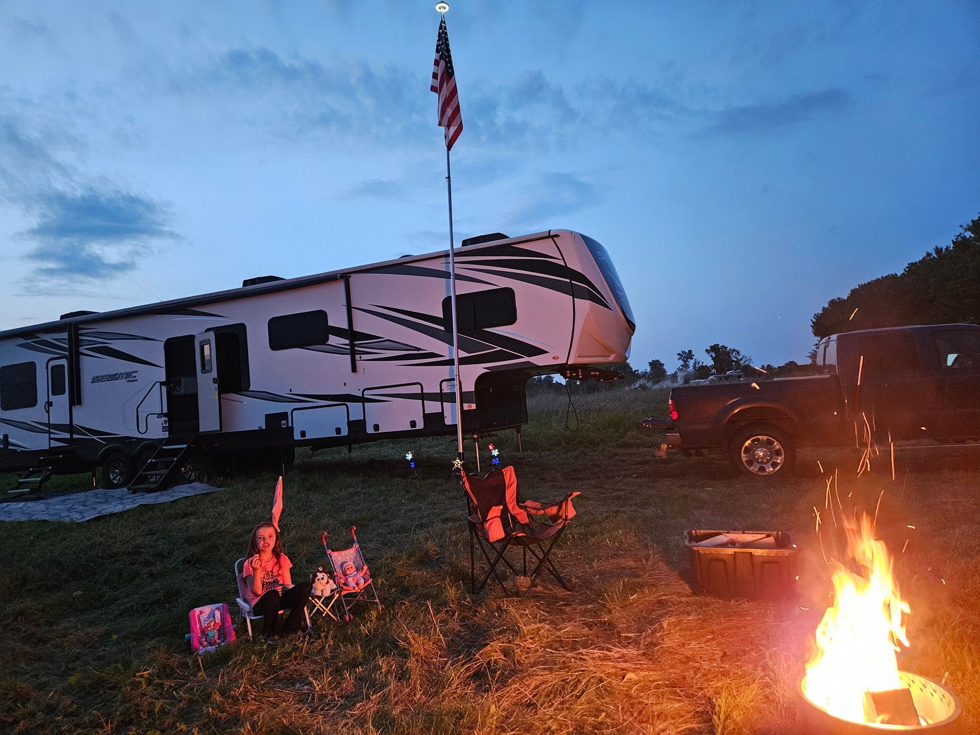 People camping by a fire beside an RV at dusk, with an American flag flying overhead.