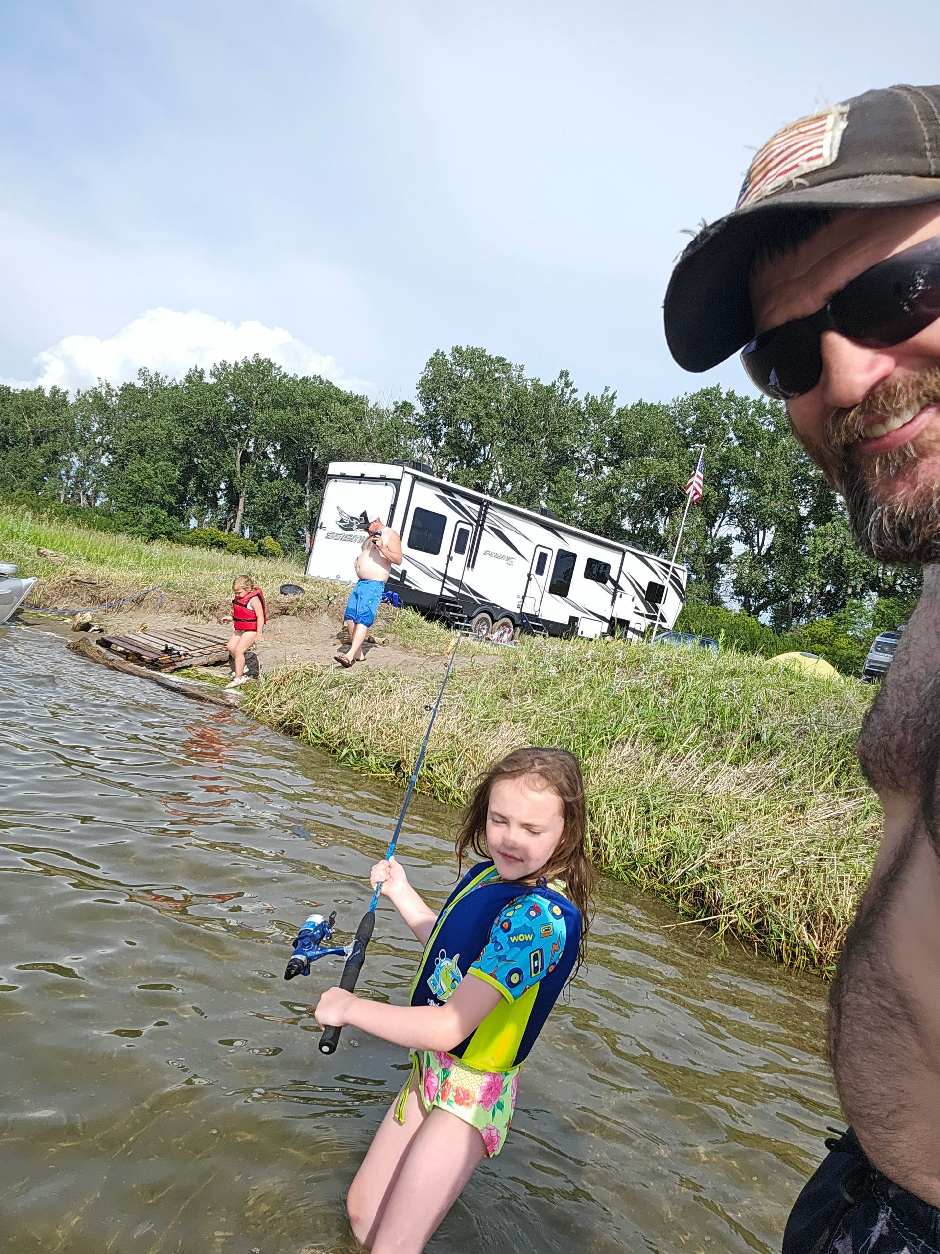 Child fishing in shallow water near a tipped RV, with two people on shore and a man in a cap smiling at the edge