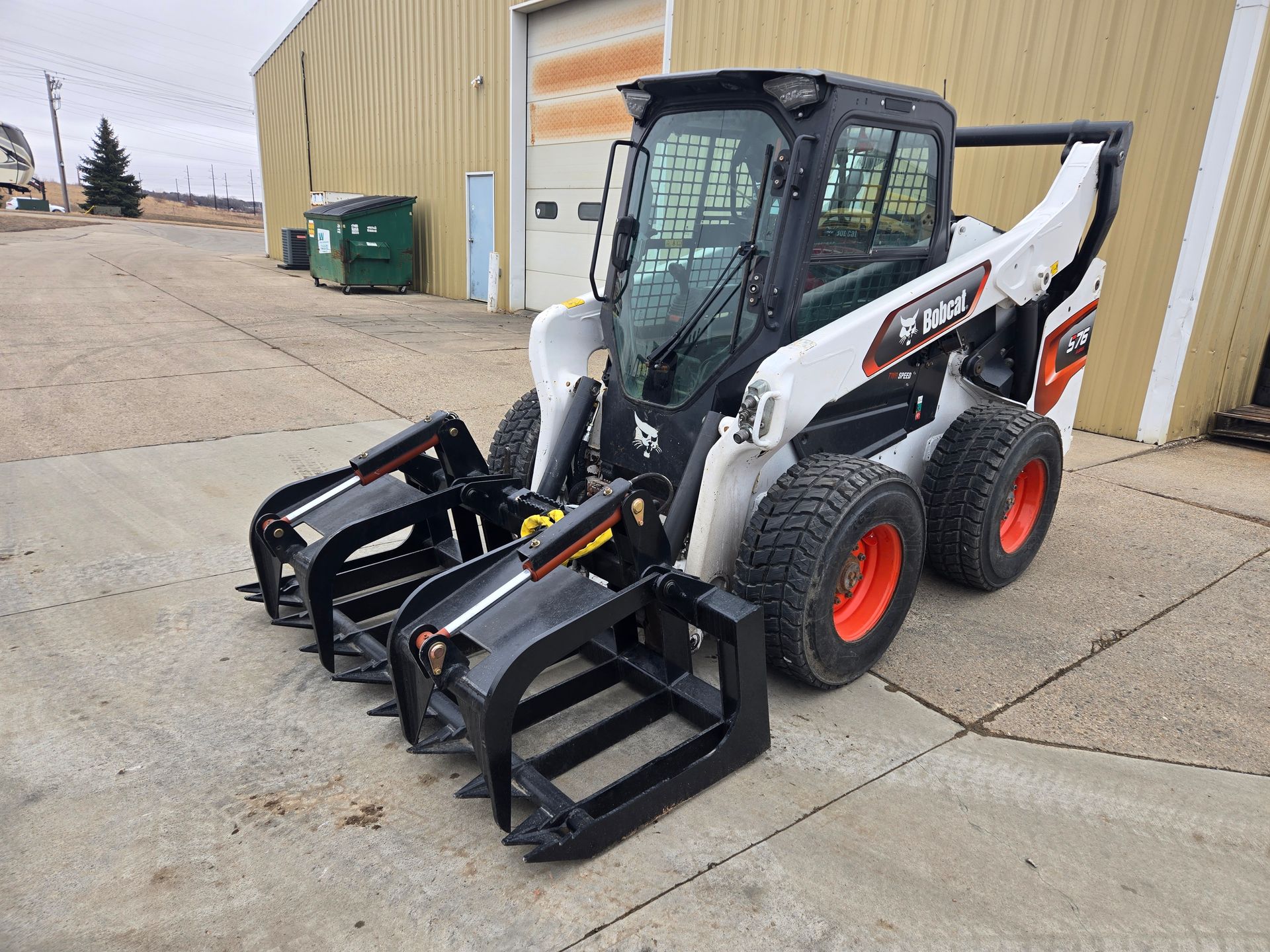 White skid-steer loader with pallet forks parked outside a beige building on a concrete lot.