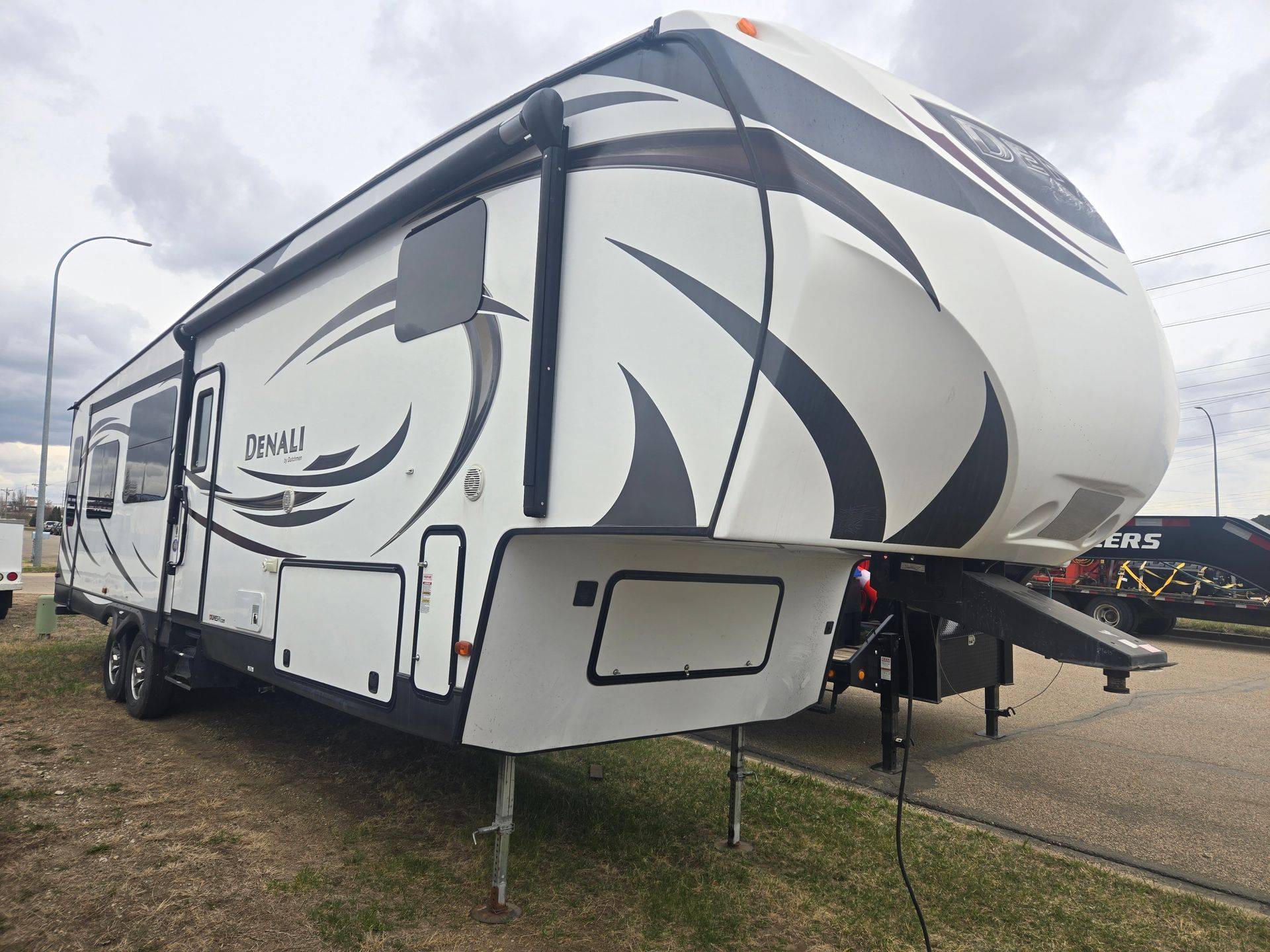 White and gray fifth-wheel RV trailer with black swoosh graphics parked on a lot under cloudy sky