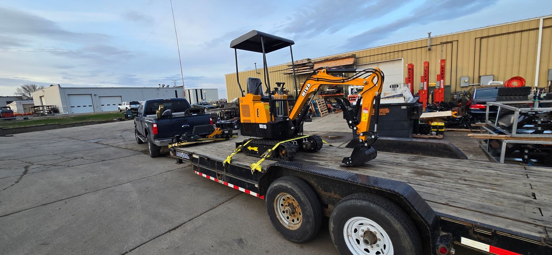Orange mini excavator on a flatbed trailer in an industrial yard under a cloudy sky.