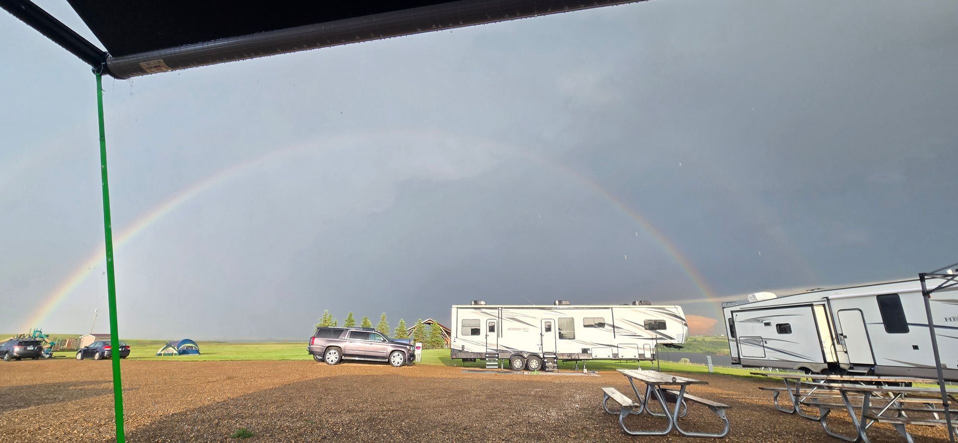 Rainbow over RVs and a parked car on a gravel lot under a cloudy sky