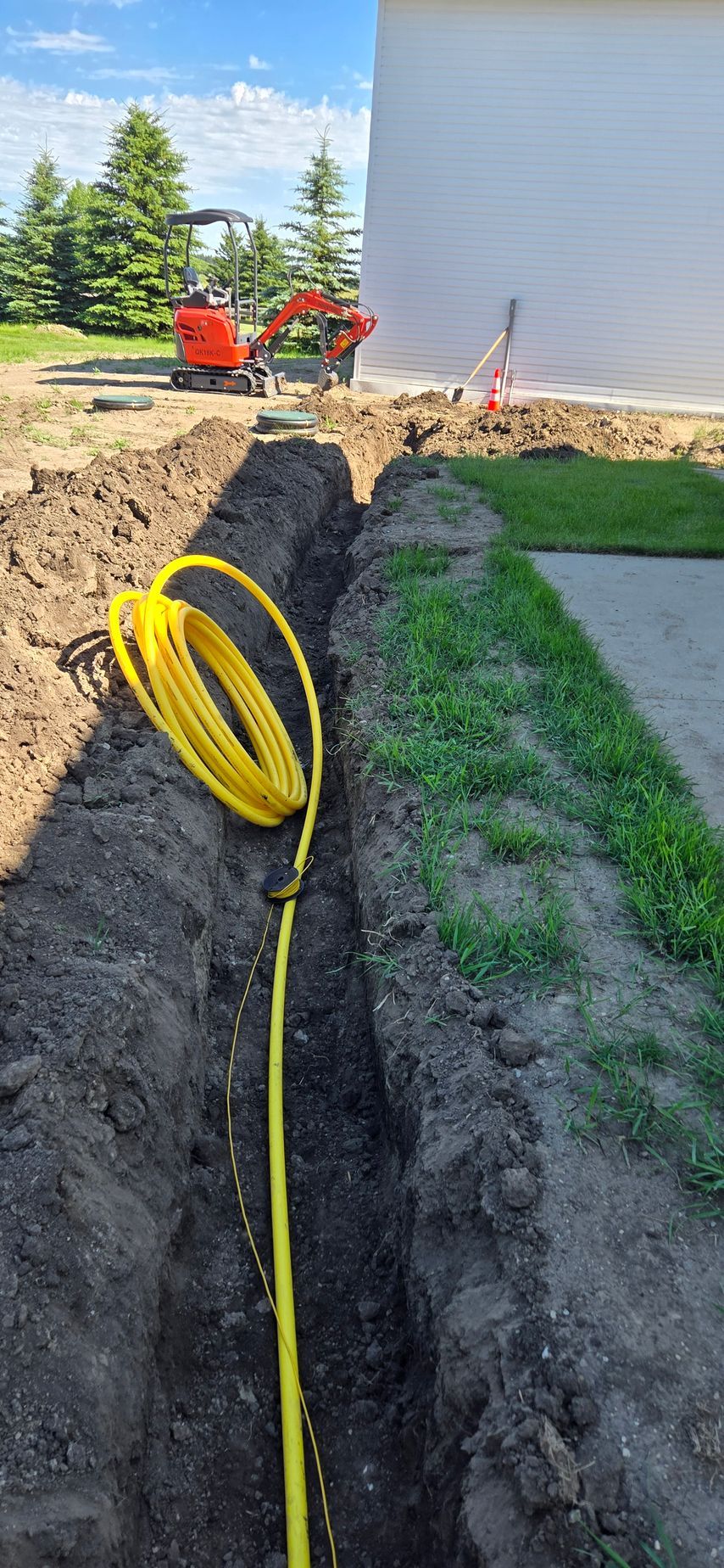 Yellow hose in a trench beside a house, with orange equipment and fresh dirt along the foundation.