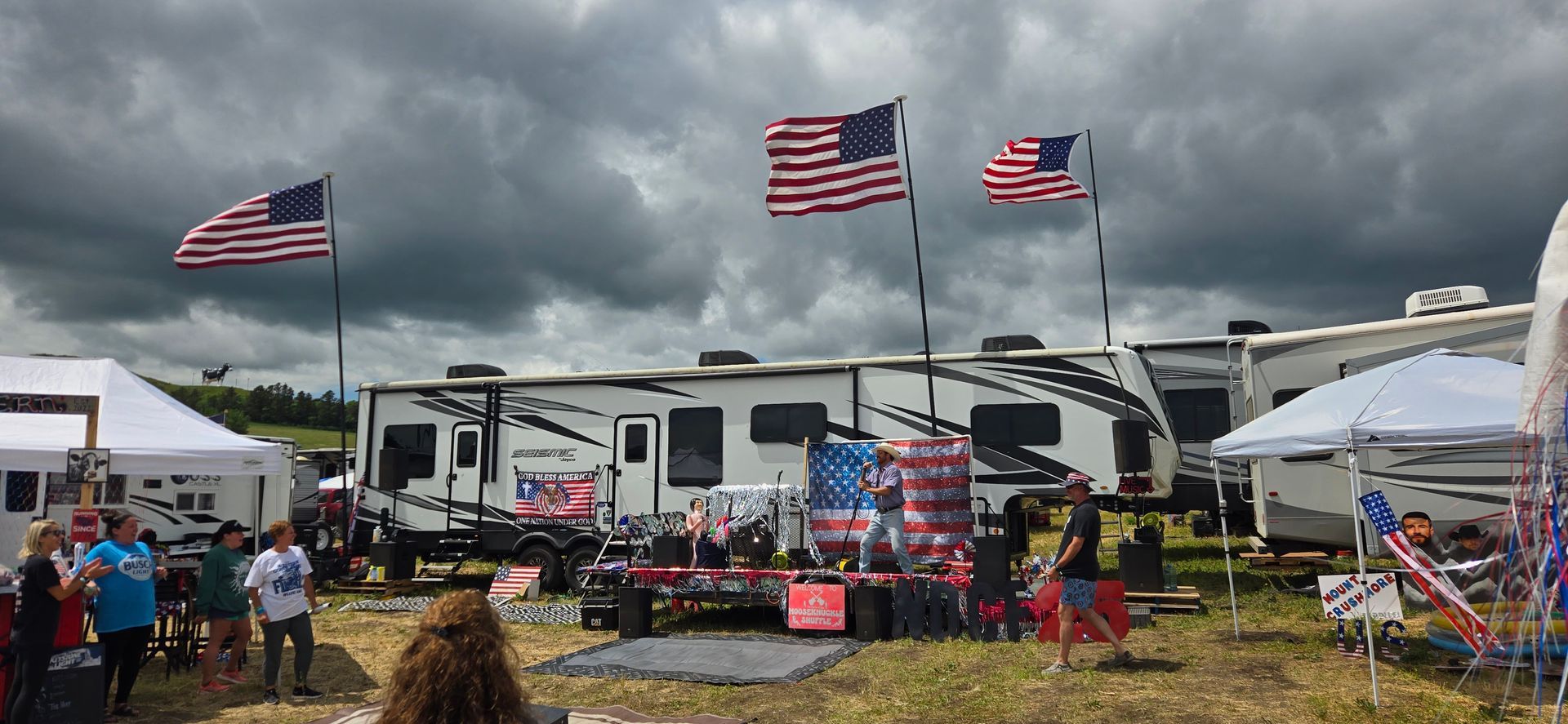 RVs and tents at an outdoor event under stormy skies, with large American flags flying overhead