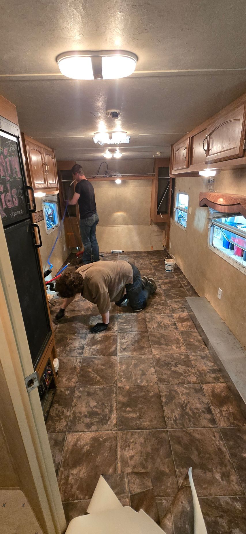 Narrow RV hallway with two people moving furniture and storage cabinets on both sides.