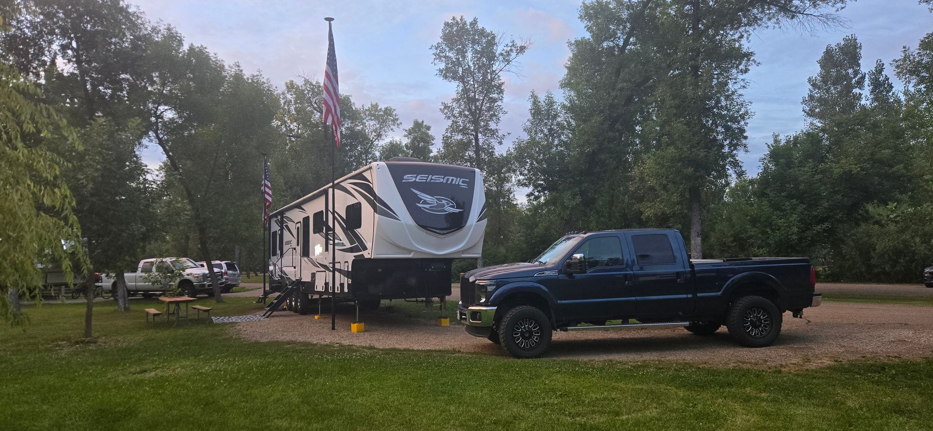 Blue pickup truck parked beside a white RV in a grassy wooded campsite