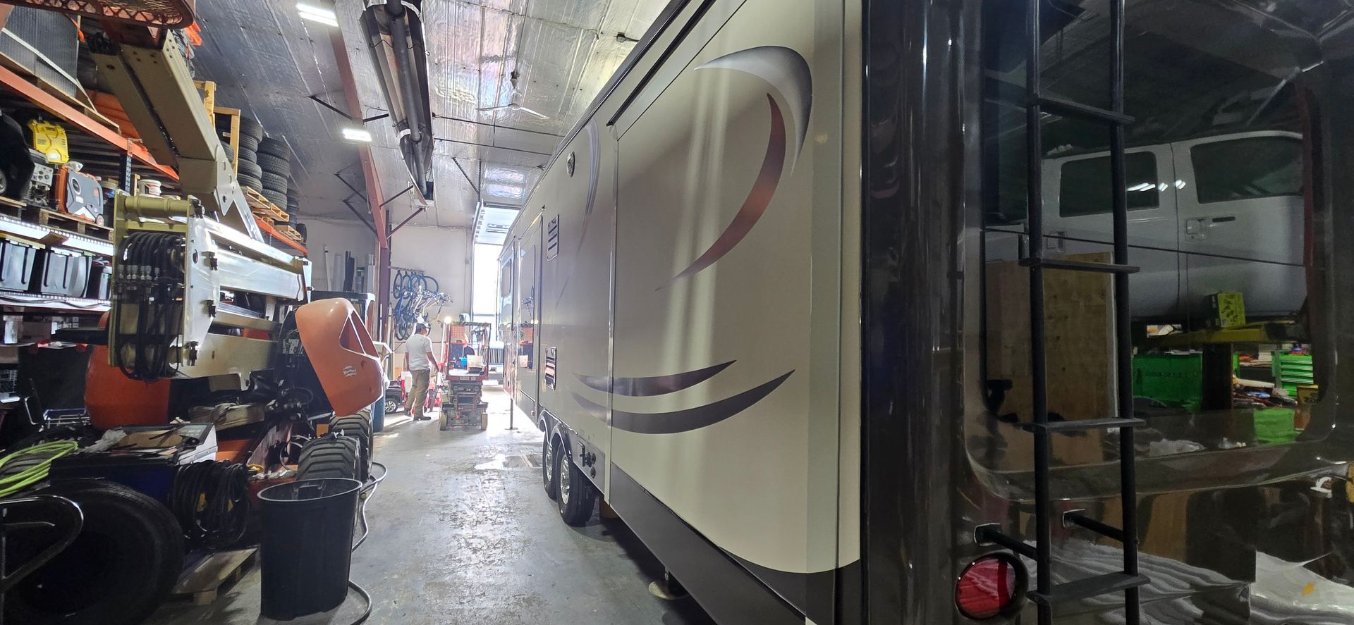 A narrow workshop aisle with a large white RV/trailer and tools, cables, and equipment on the left.