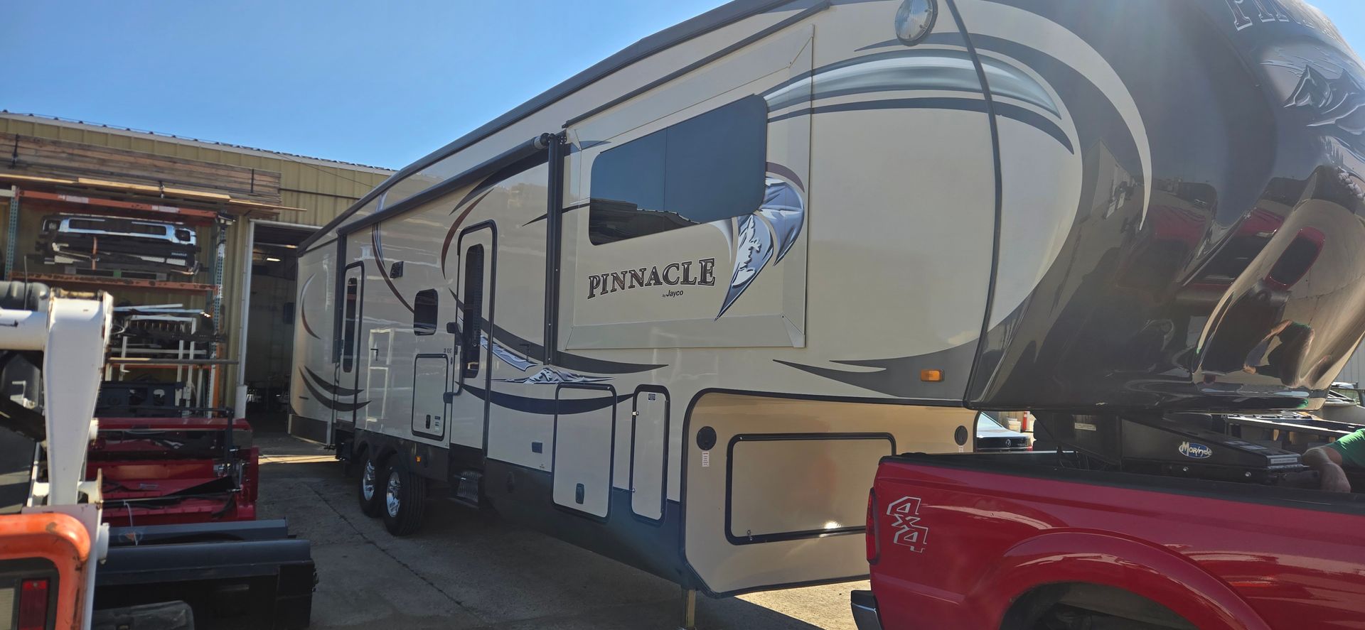 Large beige and tan travel trailer parked beside red vehicles in a lot