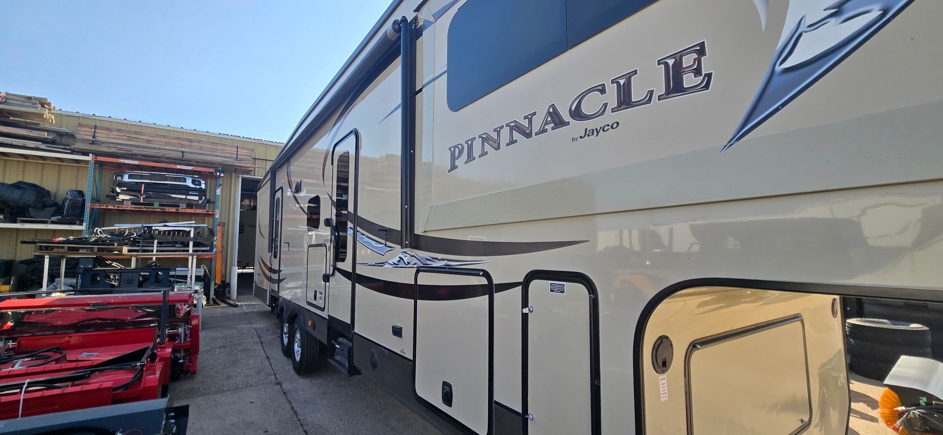 White Pinnacle RV parked beside a red truck in a storage lot under a blue sky
