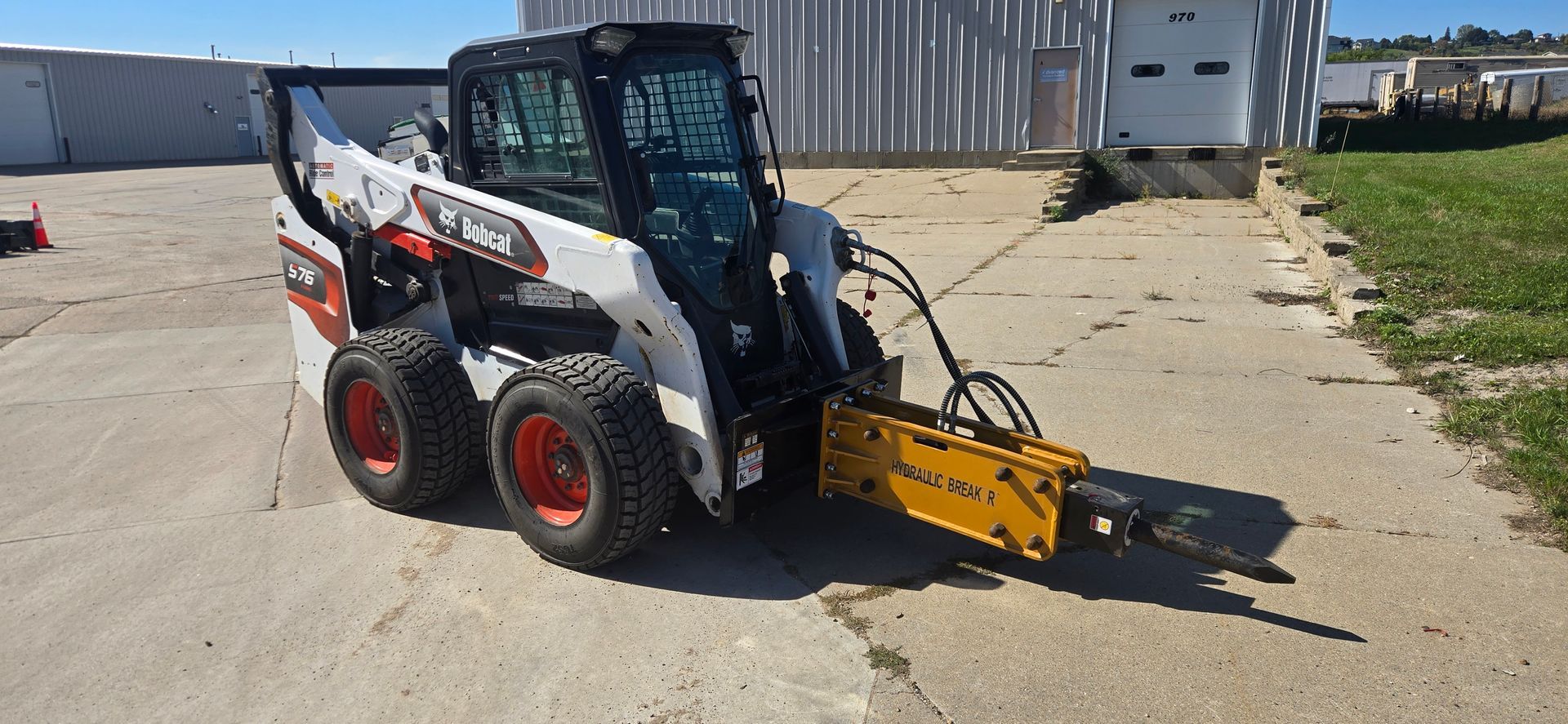 White Bobcat skid steer with yellow attachment parked on concrete near a building and grassy edge