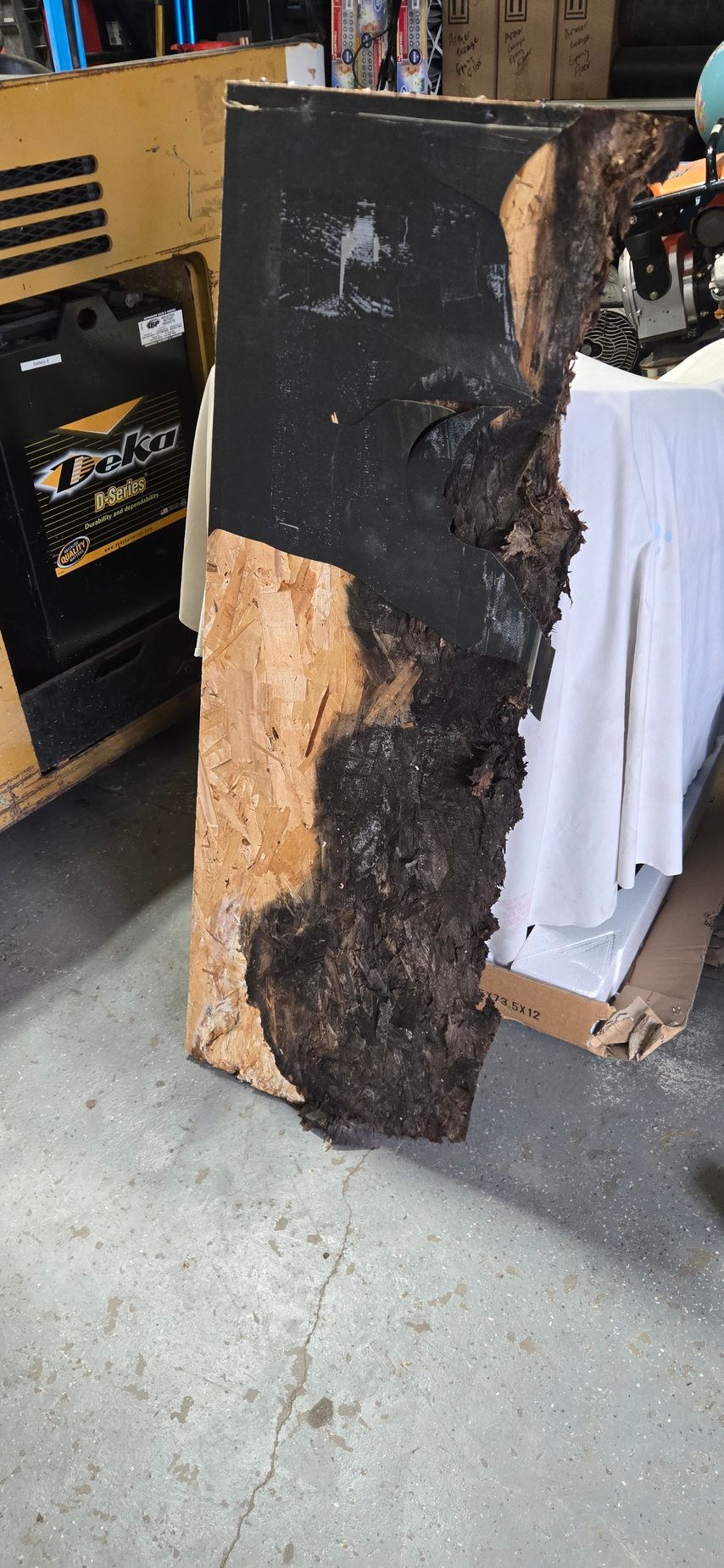 Charred wooden board leaning in a garage, with a blackened top and burnt, cracked lower half.