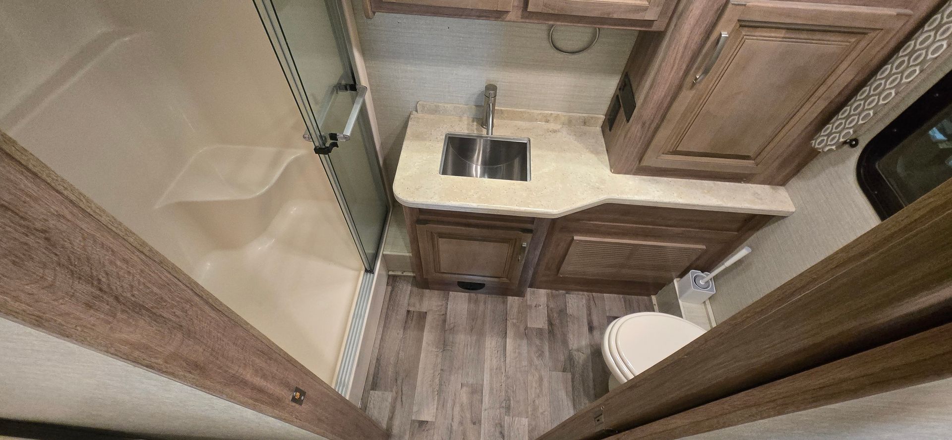 Compact RV bathroom with sink, wooden cabinets, toilet, and glass shower door