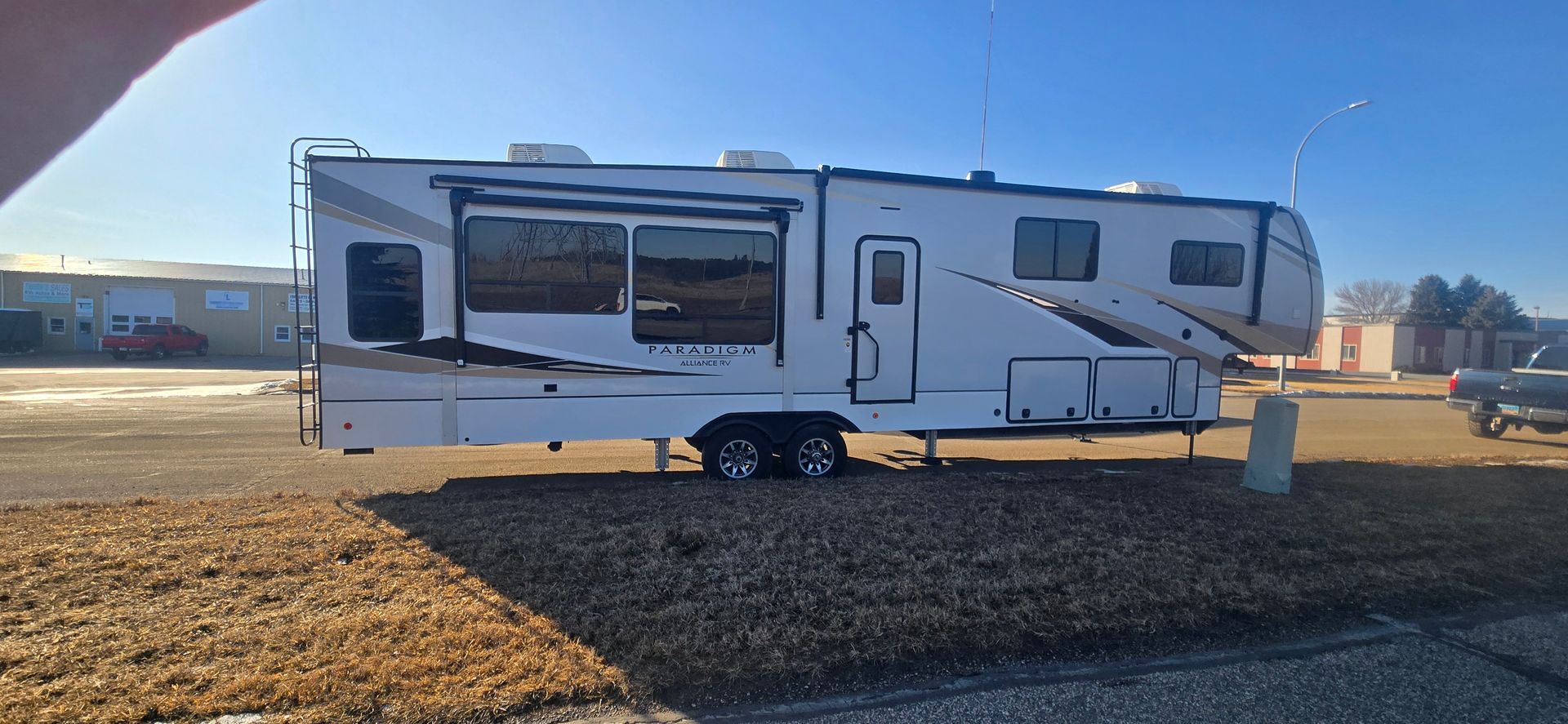 White travel trailer parked on a gravel lot in bright sunlight, with awnings extended.