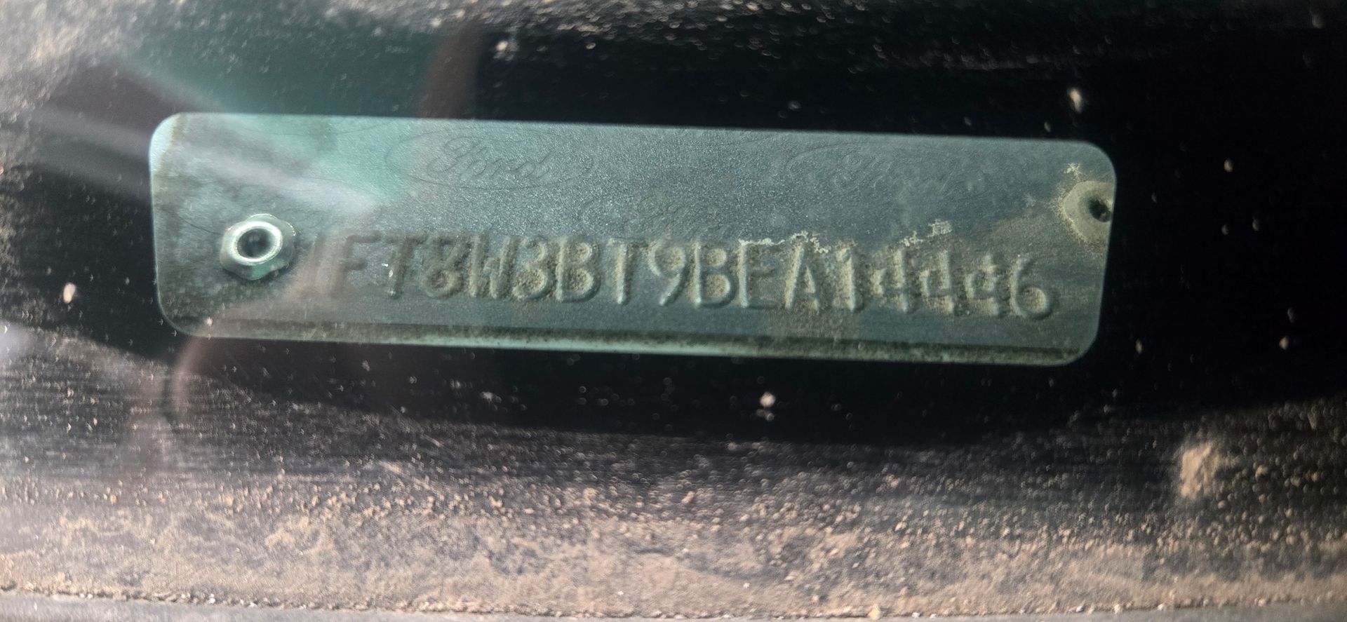 Close-up of a weathered metal identification plate with stamped alphanumeric text on a dirty surface.