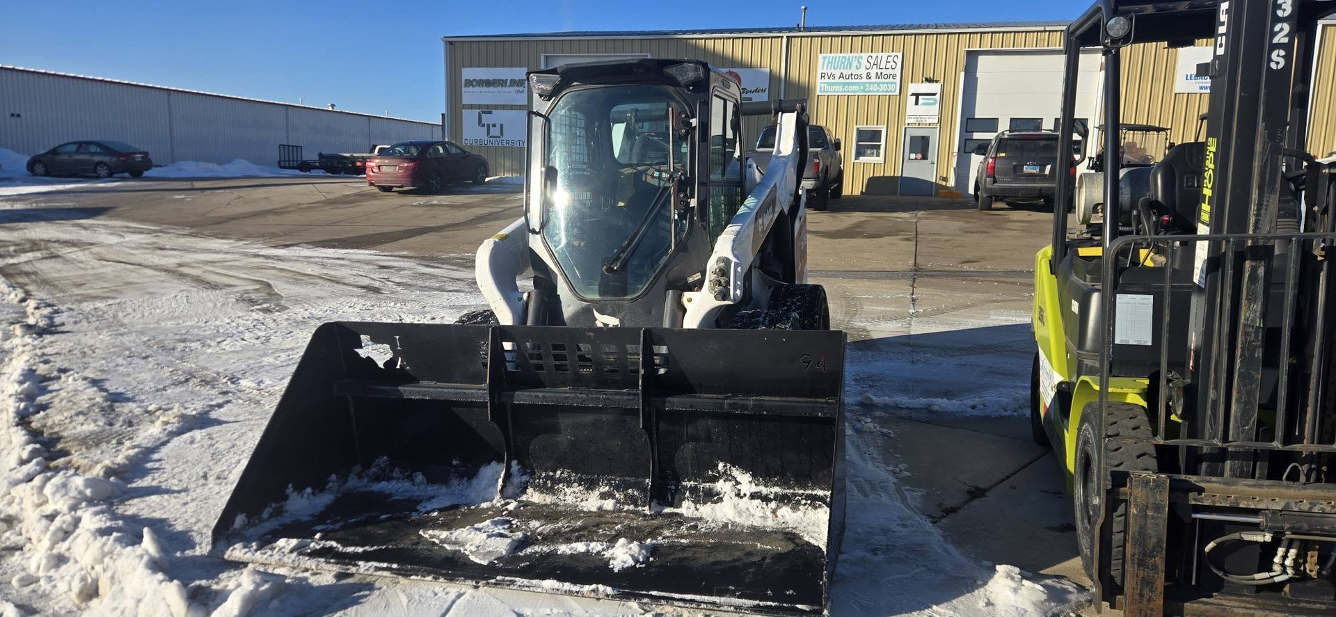 Front loader with raised bucket in a snowy industrial yard beside a forklift