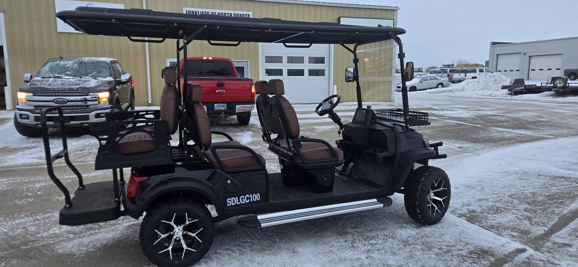 Black off-road golf cart with roof and gear parked on a snowy lot beside a gray truck
